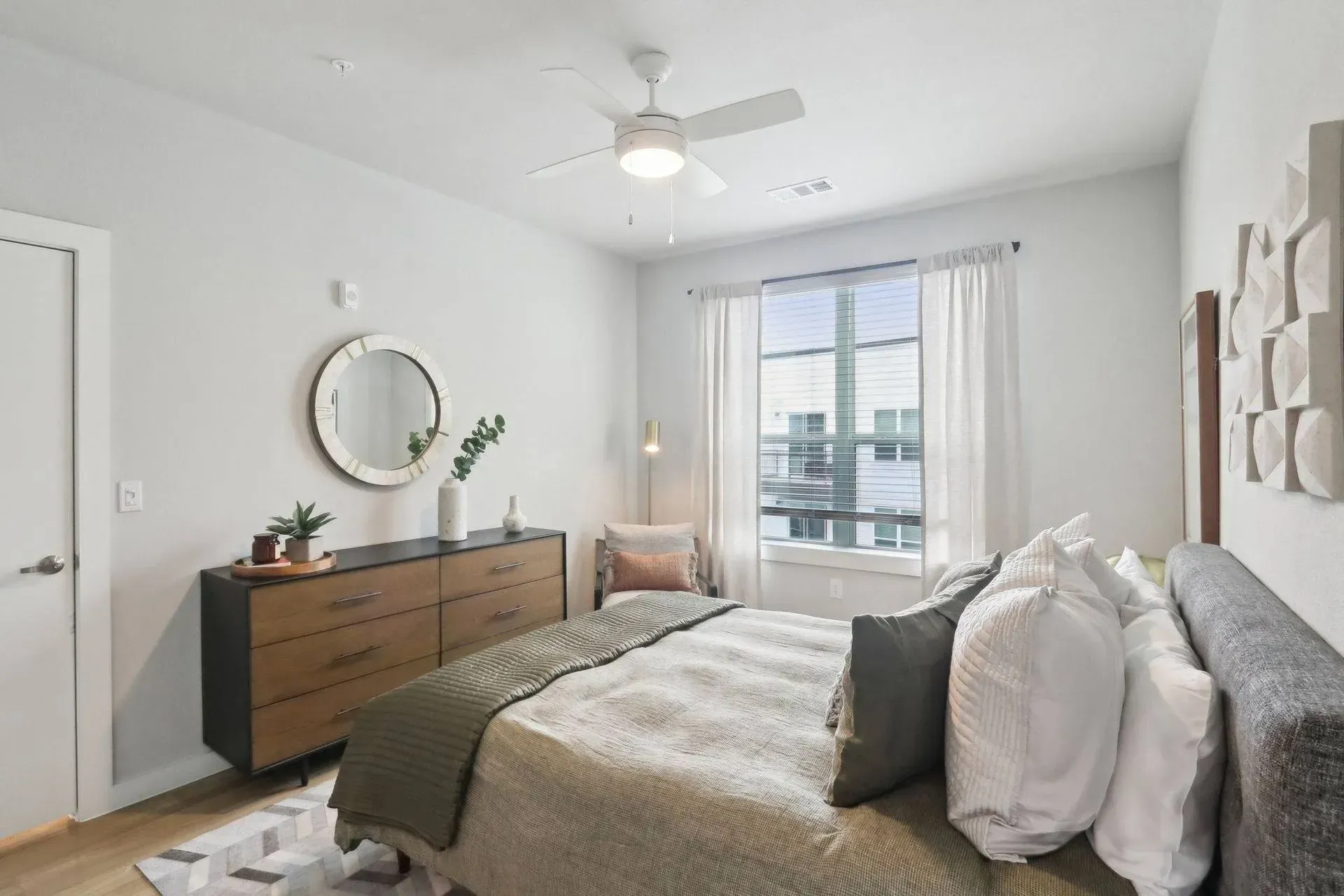 Bedroom with bed, dresser, mirror, window, and ceiling fan. Light gray walls, neutral tones.