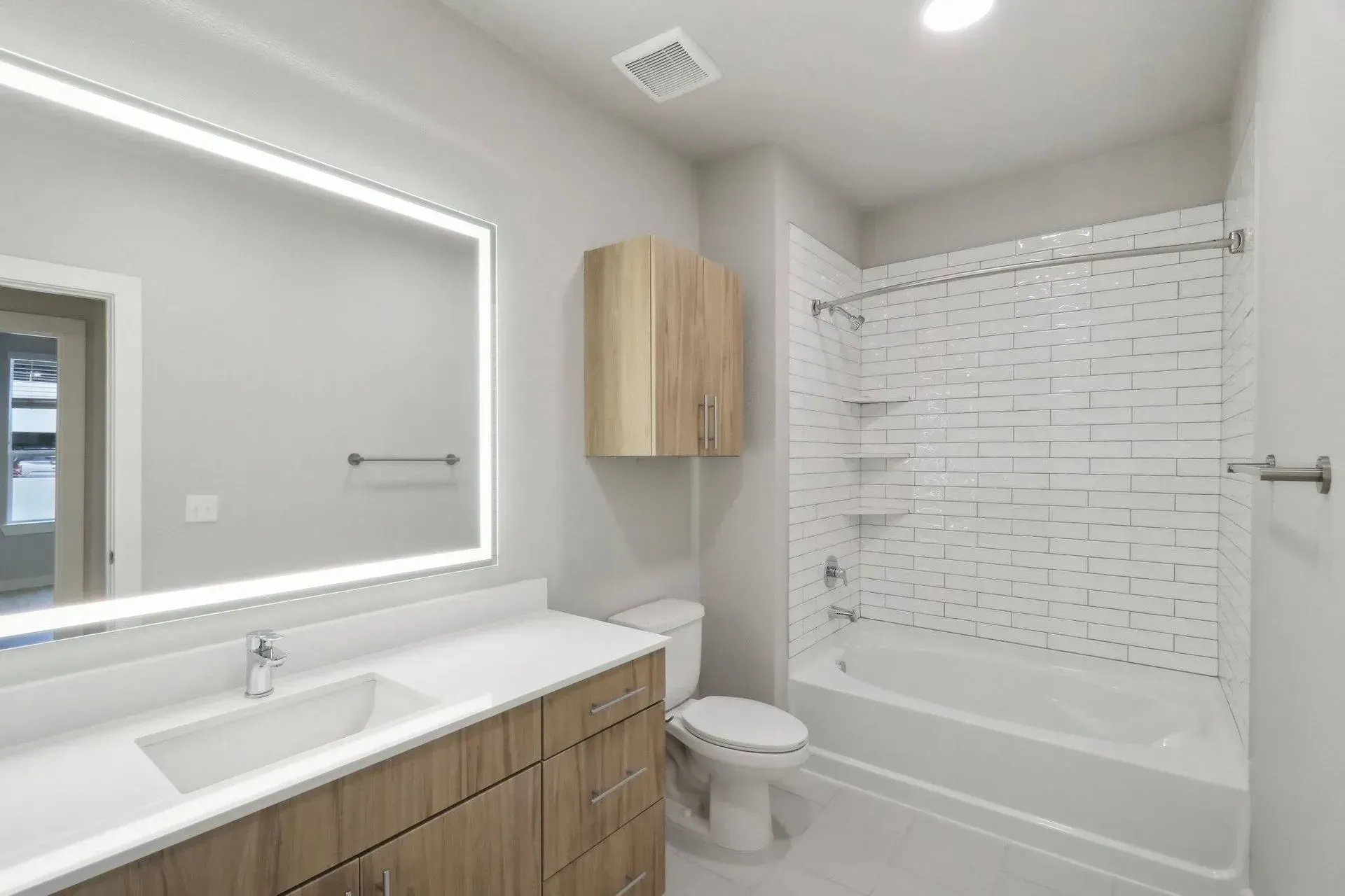 Modern bathroom with white walls, light wood vanity and cabinet, large mirror with light, and a white tile shower.