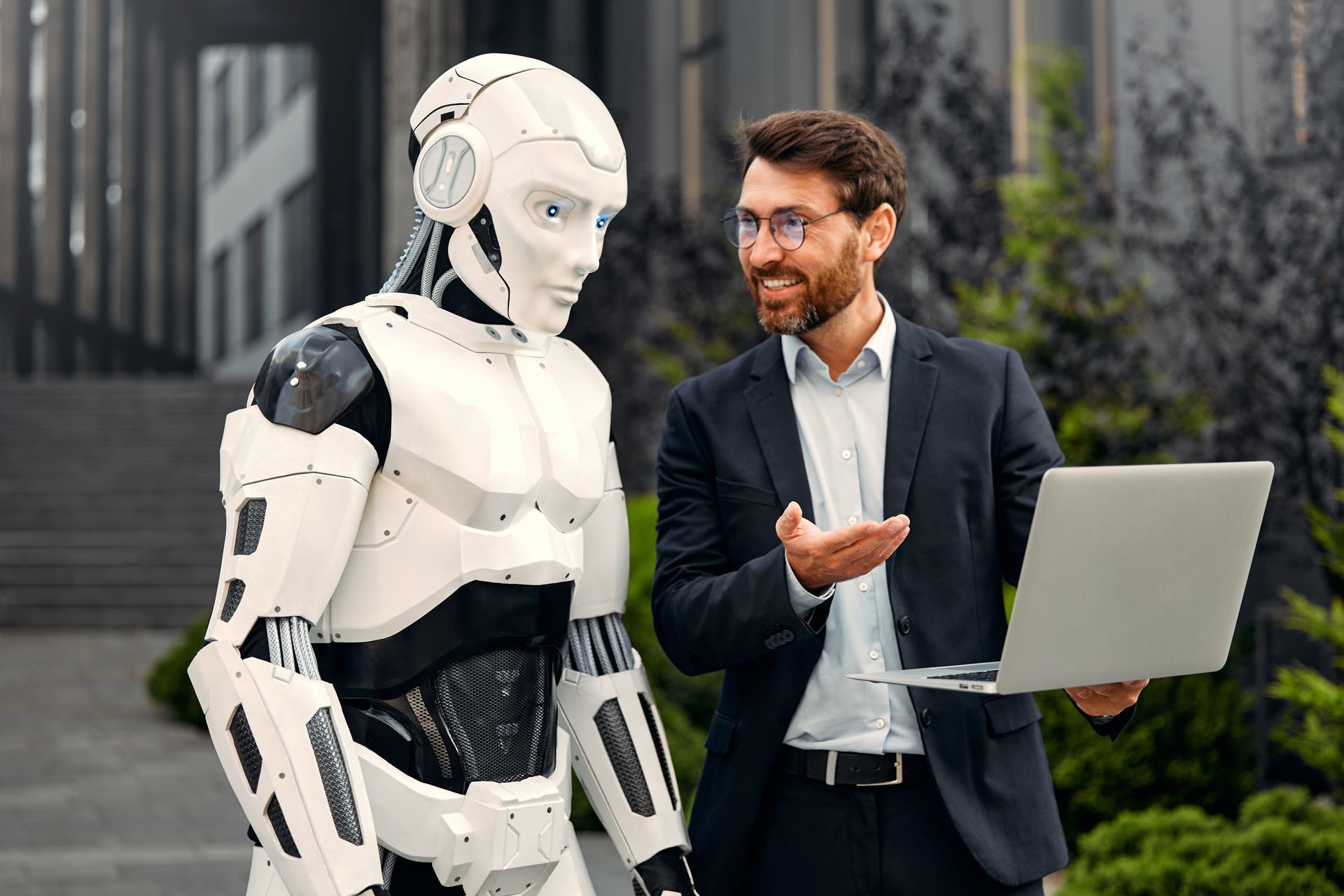Een man in een pak praat met een robot terwijl hij een laptop vasthoudt.