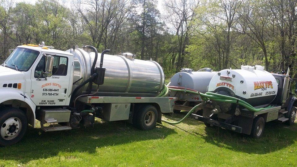 Septic trucks parked on grass, hoses connected. White and silver tanks.