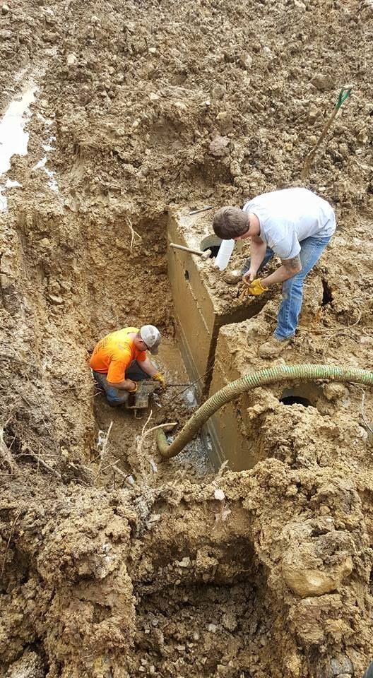 Two workers emptying a tank in a muddy excavation. One kneels with a tool, the other pours from a hose.