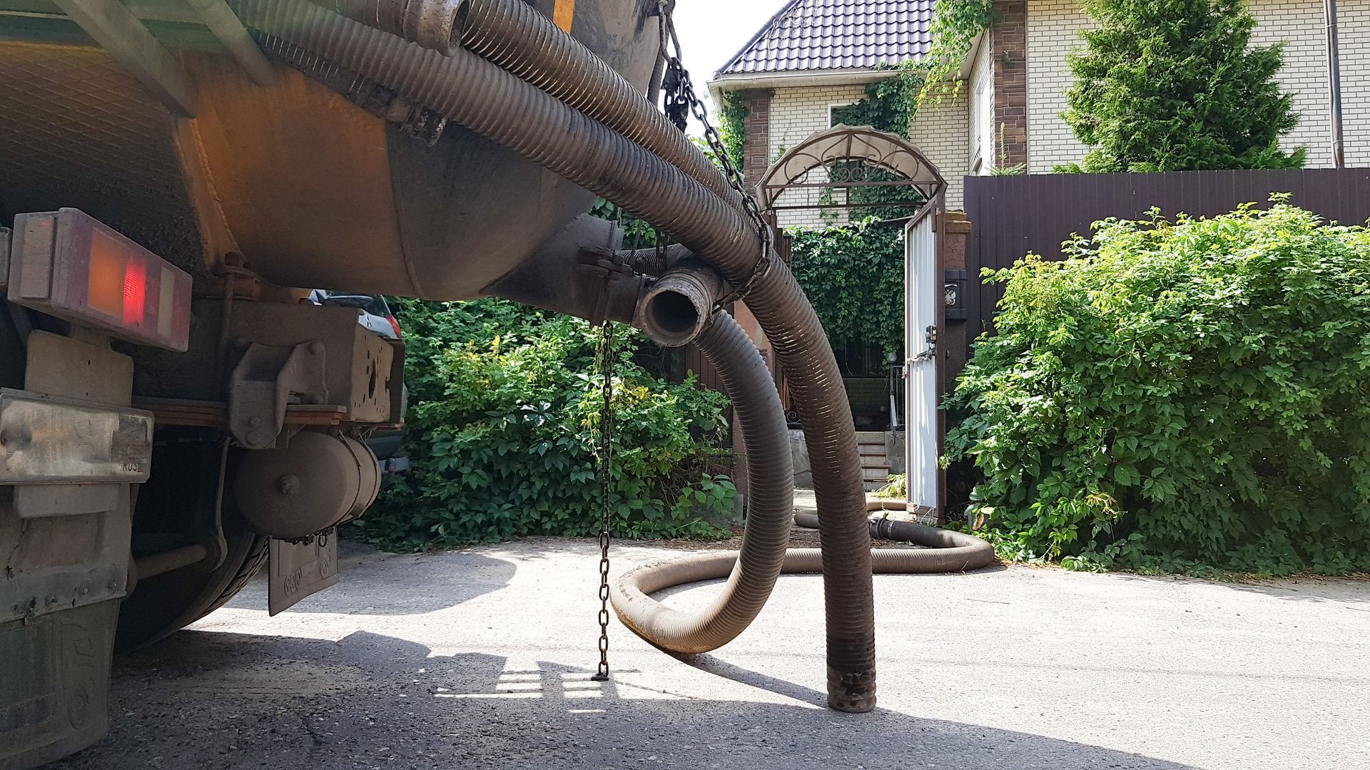 A sewage truck is parked on a paved driveway with hoses extended, near a house and garden.
