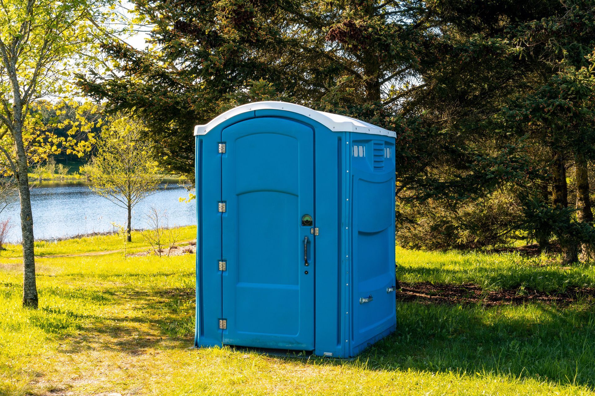 Blue portable toilet in a grassy area with trees and a lake in the background.