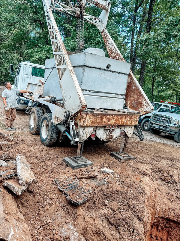 A large truck with a drilling rig is positioned over an excavated hole in a dirt area. A person stands nearby.