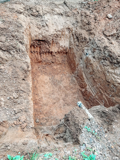 A rectangular trench dug into earthen soil, revealing layers of brown and orange sediment.