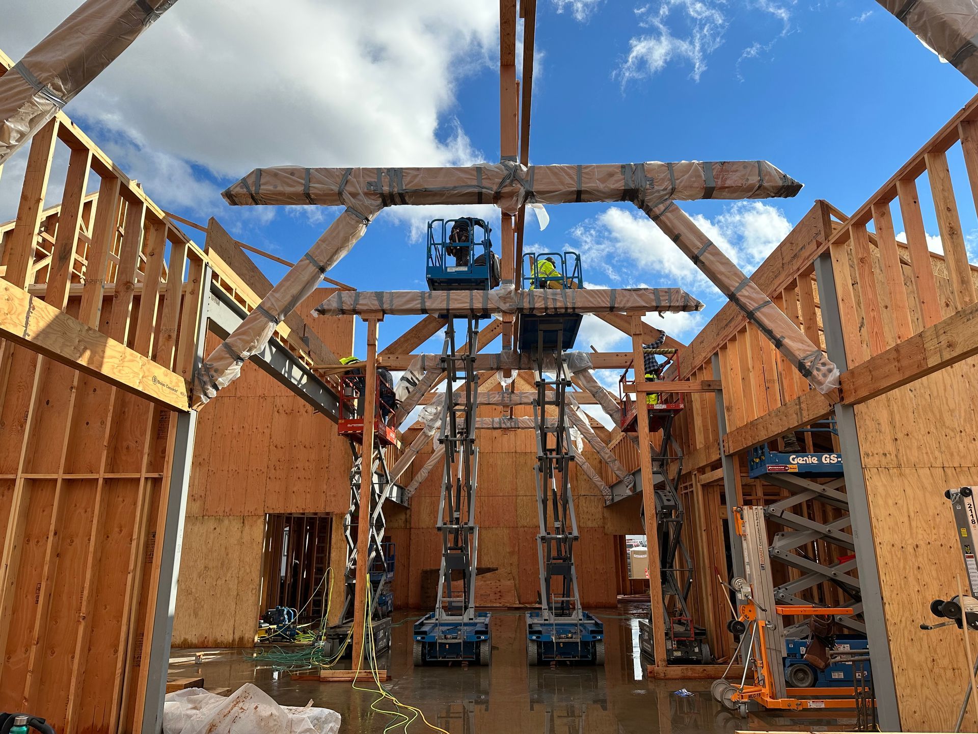 Construction site: workers on lifts installing wooden beams for a building frame, under a blue sky.