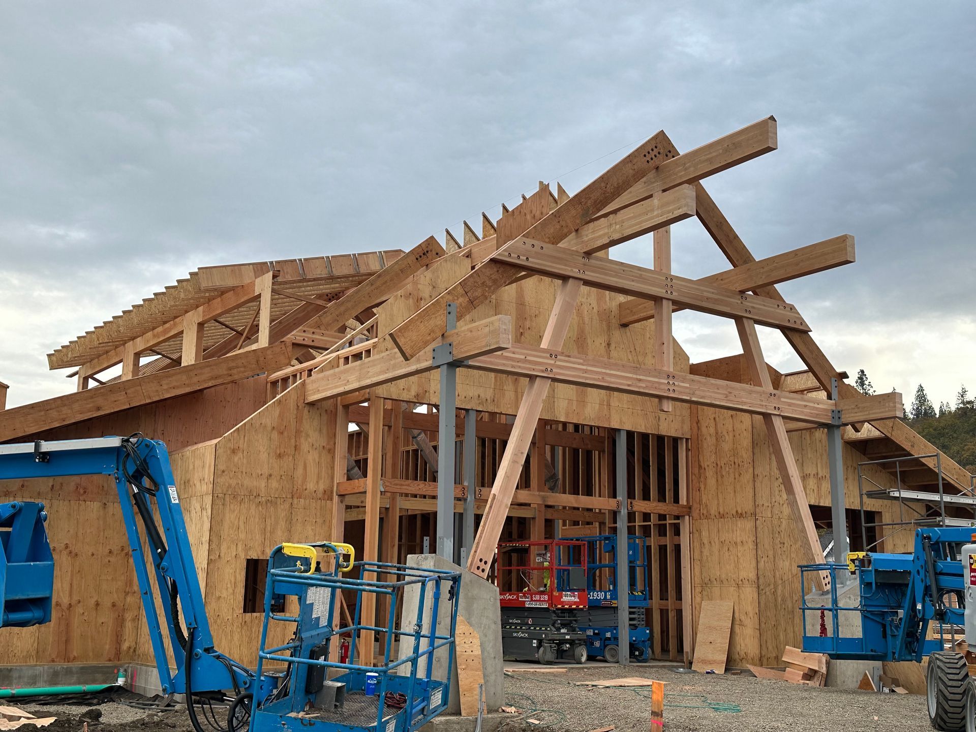 Building under construction; wood framing, scaffolding, and blue lift equipment against cloudy sky.