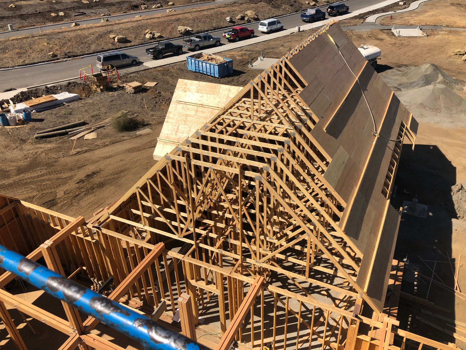 Construction site with wooden roof trusses being built. Blue pipe, cars and dirt are visible.