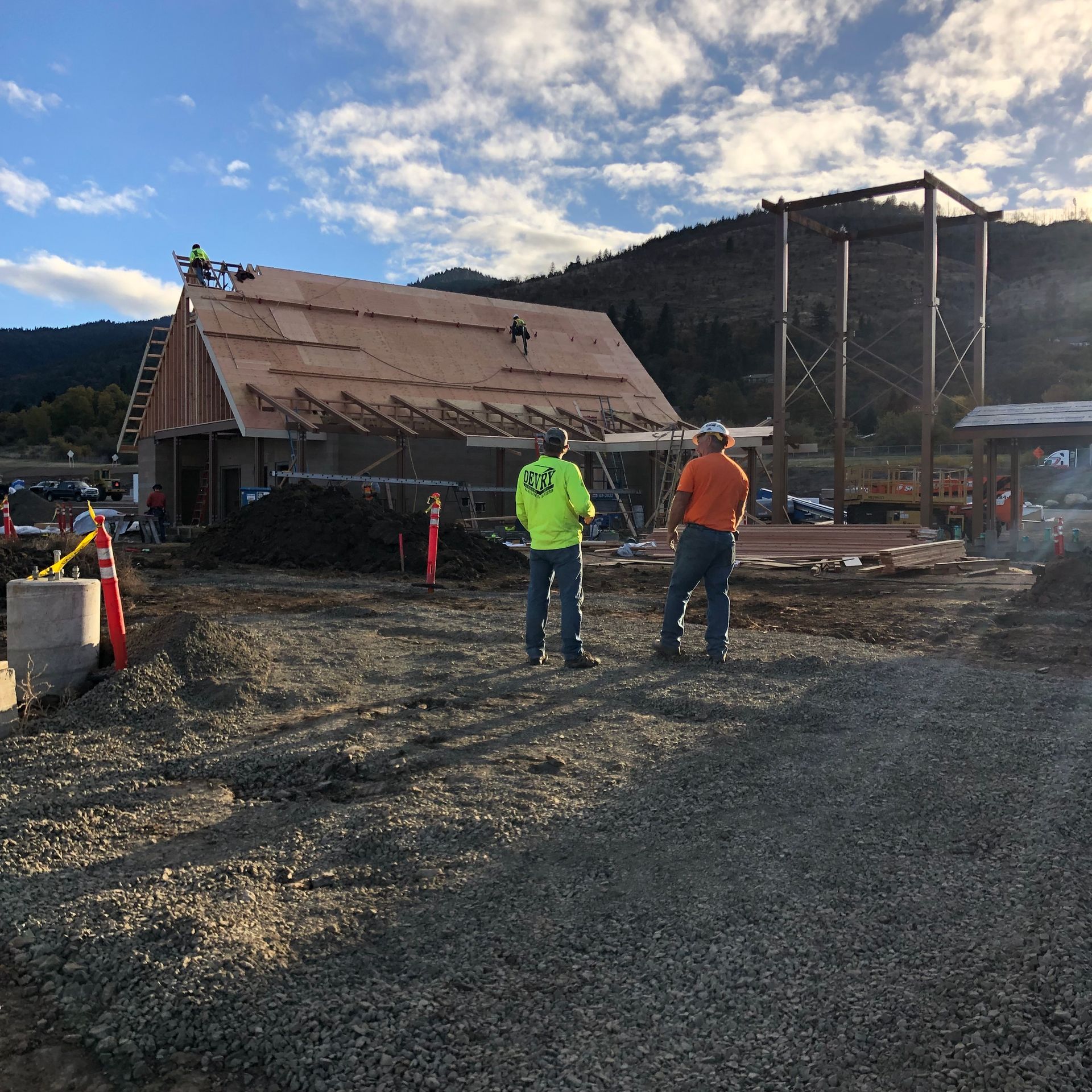 Construction workers on a roof, building a wooden structure on a gravel lot with a mountain backdrop.