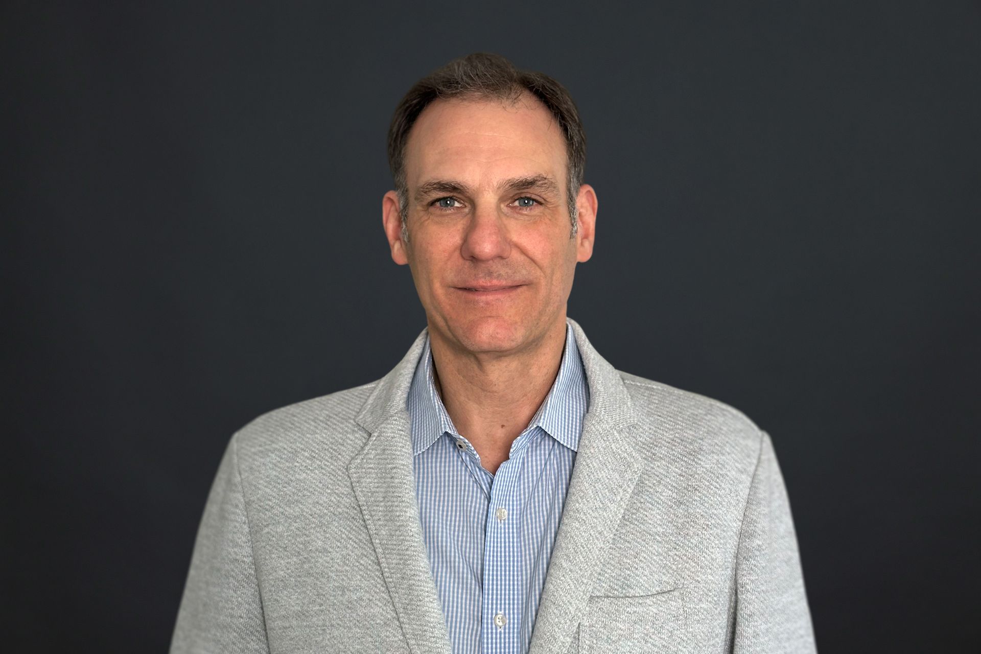 A man wearing a gray jacket and a blue plaid shirt is standing in front of a black background.