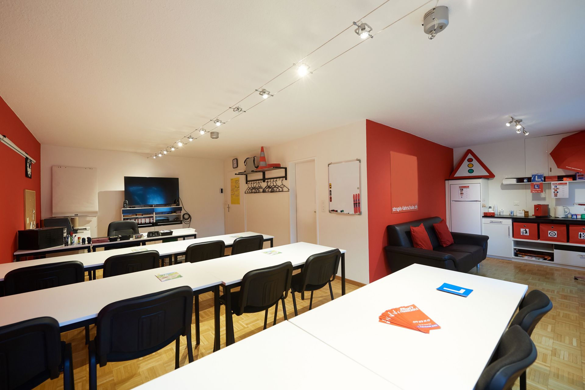 Leeres Klassenzimmer mit weissen Tischen, schwarzen Stühlen, roten Akzentwänden und einem Fernseher vorne.