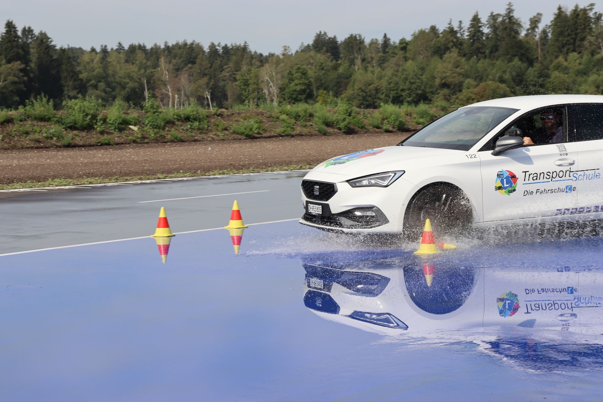Ein weisses Auto fährt über eine blaue Wasserspritzstrecke neben Verkehrskegeln und Waldkulisse.