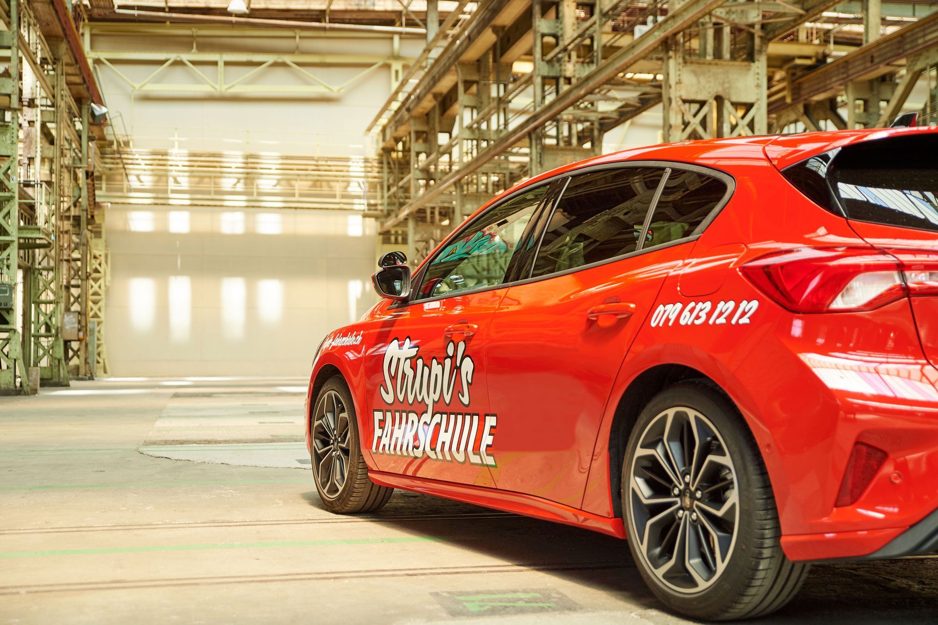 Rotes Fahrschulauto in einer hell erleuchteten Industriehalle, von hinten gesehen.