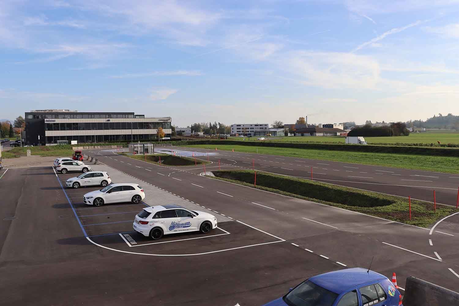 Eine Reihe weisser Fahrschulautos geparkt auf einem gepflasterten Parkplatz neben einem modernen Gebäude und einem Rasenfeld.