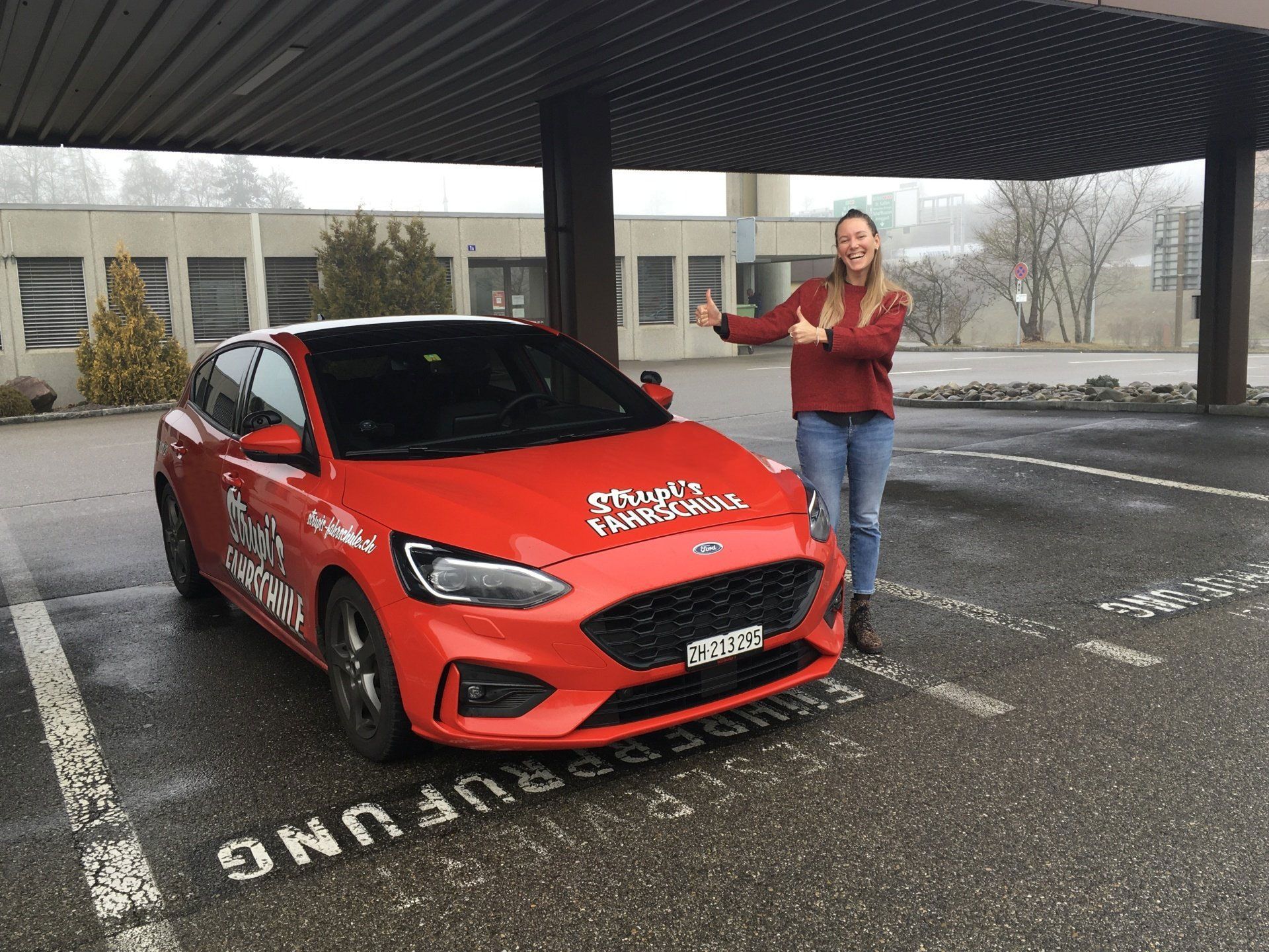 Eine Person neben einem roten Auto unter einem Dach an einem regnerischen Tag