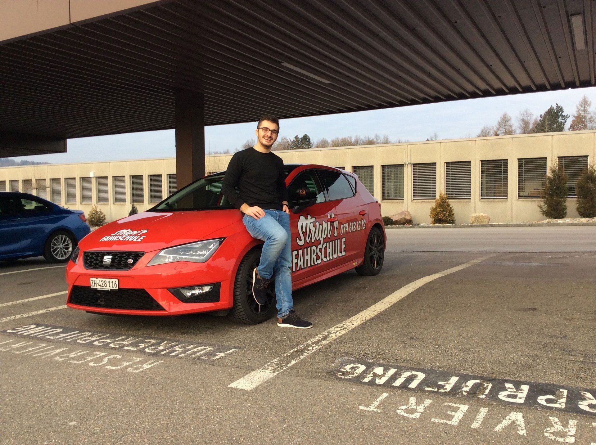 Ein Mann lehnt unter einem überdachten Parkplatz an einem roten Auto.