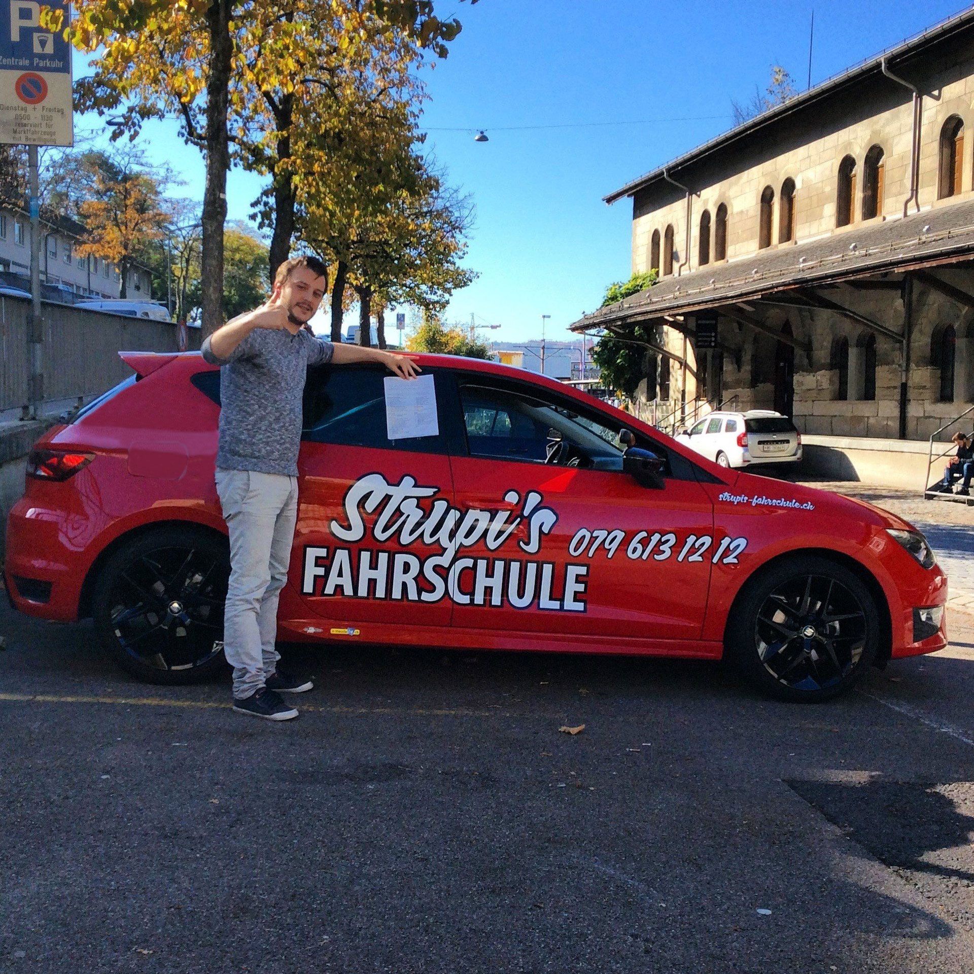 Eine Person steht neben einem roten Fahrschulwagen mit der Aufschrift „Strupis Fahrschule“ an der Seite, der auf einem Parkplatz steht.