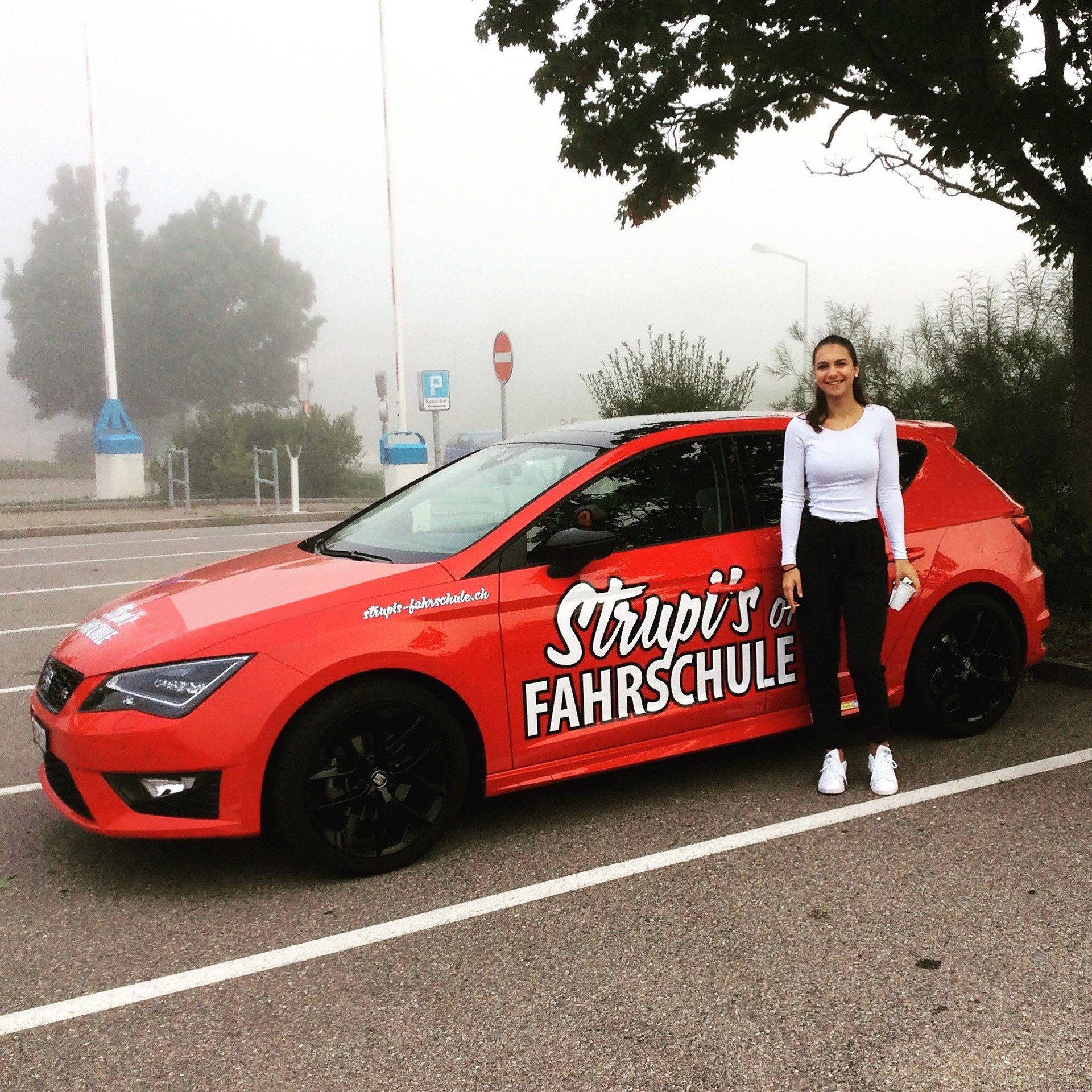 Frau steht neben einem roten Fahrschulwagen mit der Aufschrift „Strupis Fahrschule“ auf der Tür auf einem nebligen Parkplatz