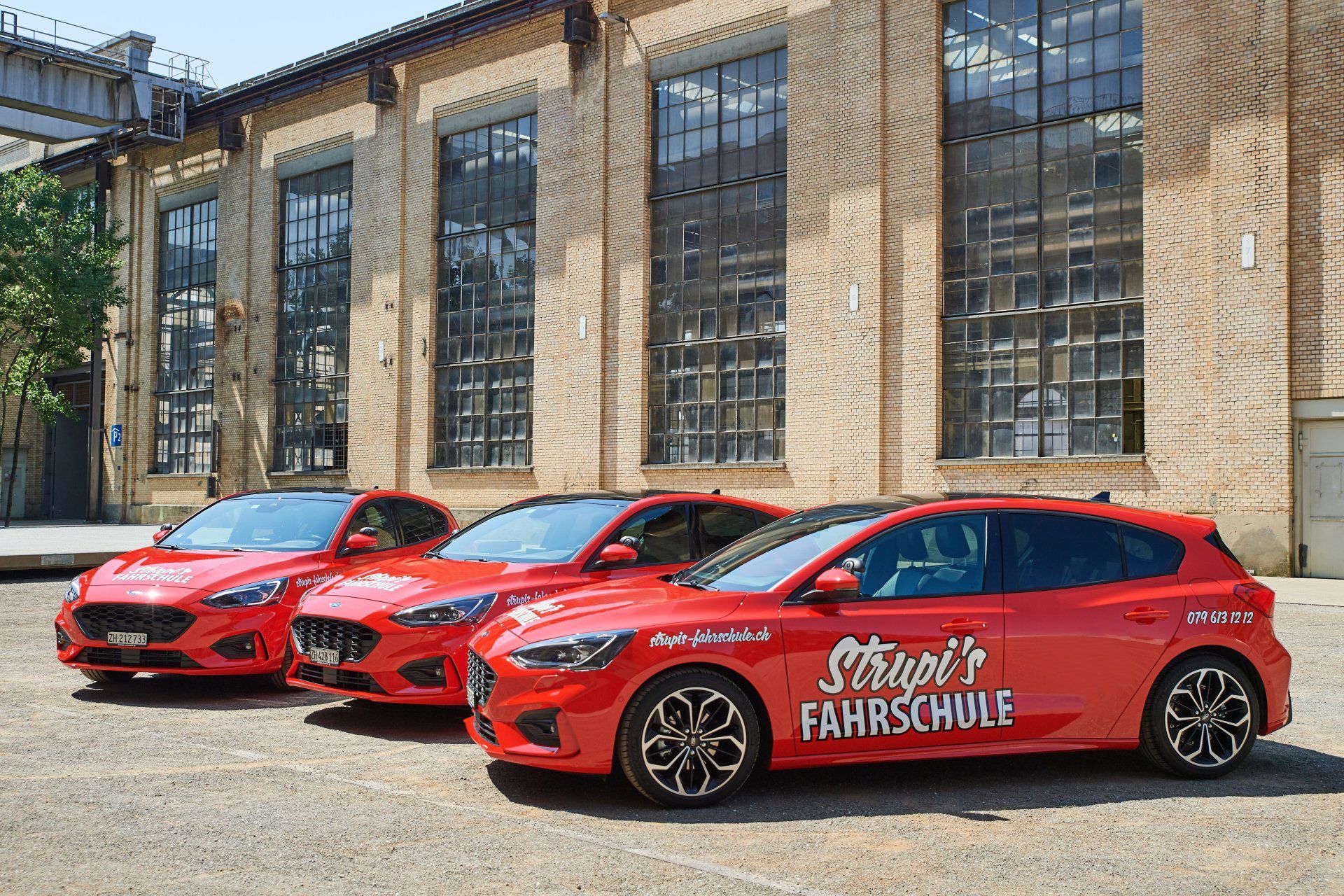 Drei rote Fahrschulwagen parkten vor einem Backsteingebäude mit hohen Fenstern.