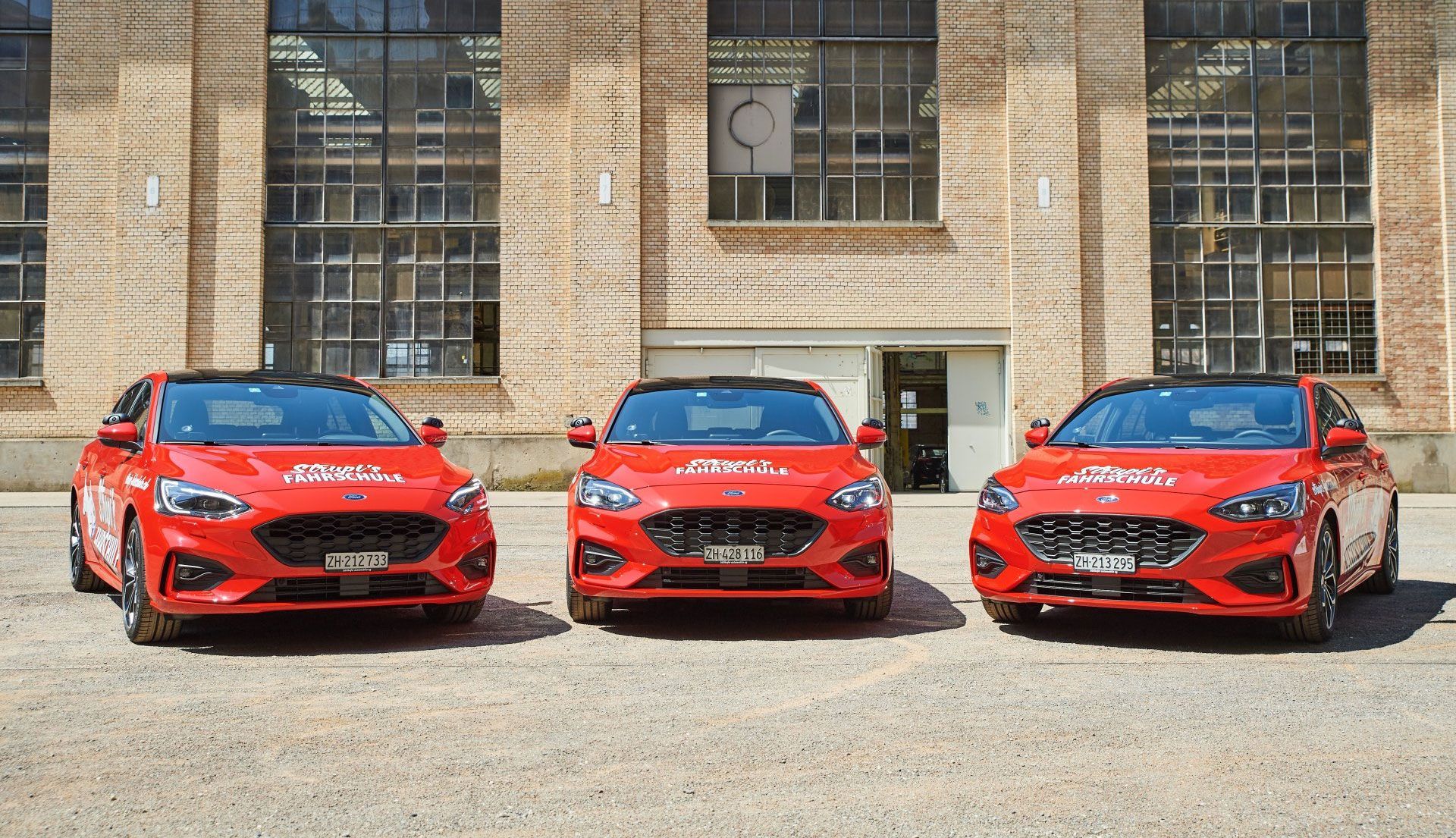 Drei rote Fahrschulautos nebeneinander vor einem Backsteingebäude geparkt.