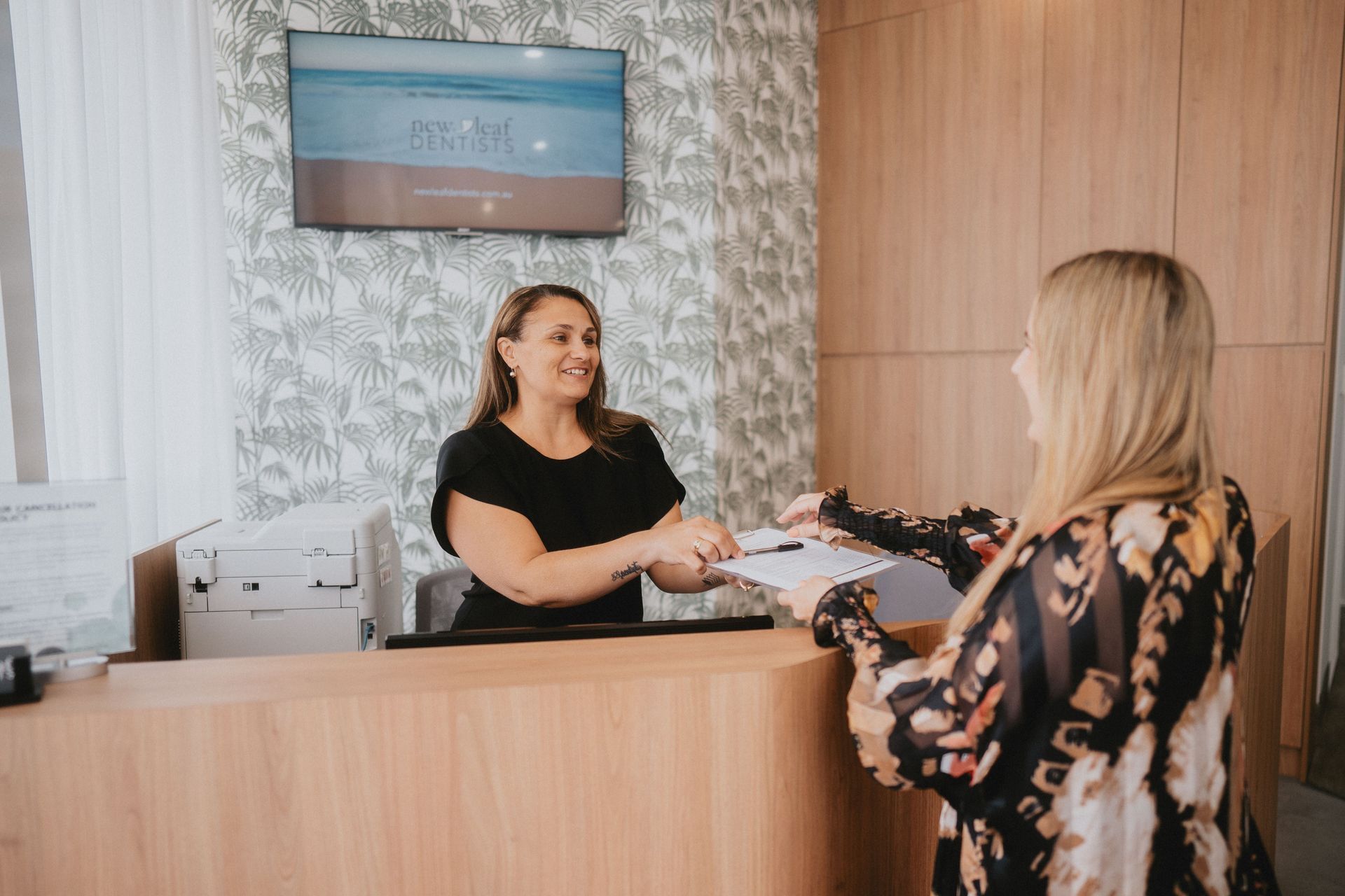 Woman at Reception Desk Handing Documents to Another Woman — New Leaf Dentists In Erina, NSW