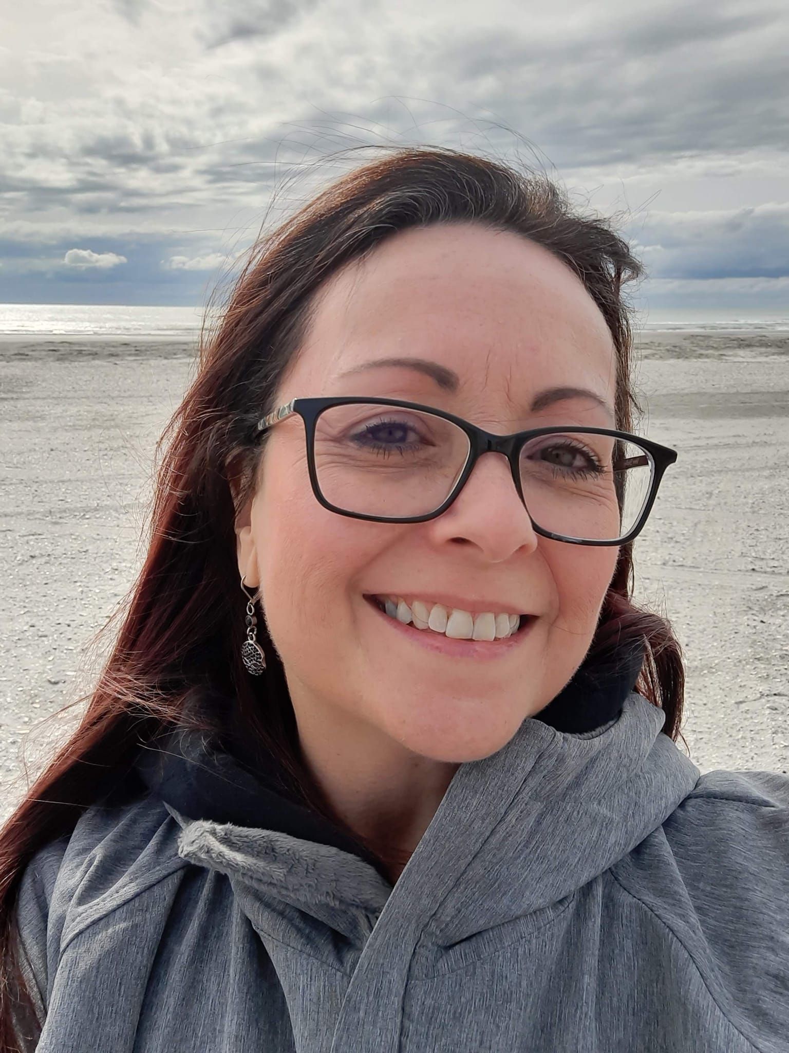 A woman wearing glasses and a gray jacket is smiling on the beach.