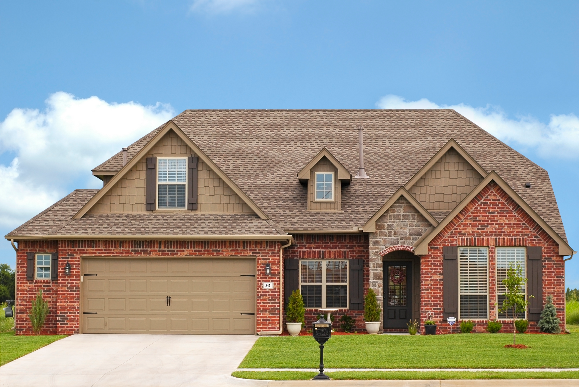 Brick house with tan garage door, shutters, and roof against a blue sky.