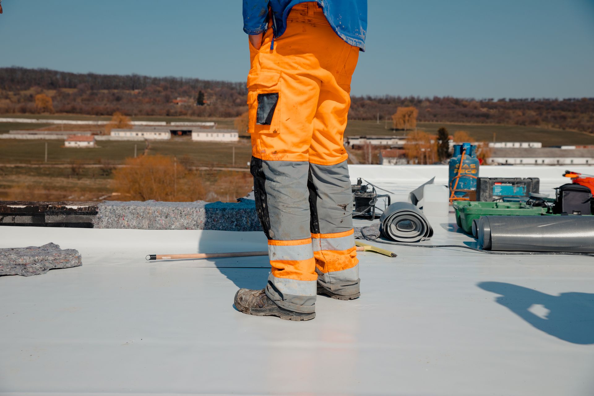 Person in orange work pants on a white roof, with tools nearby and a building in the background.