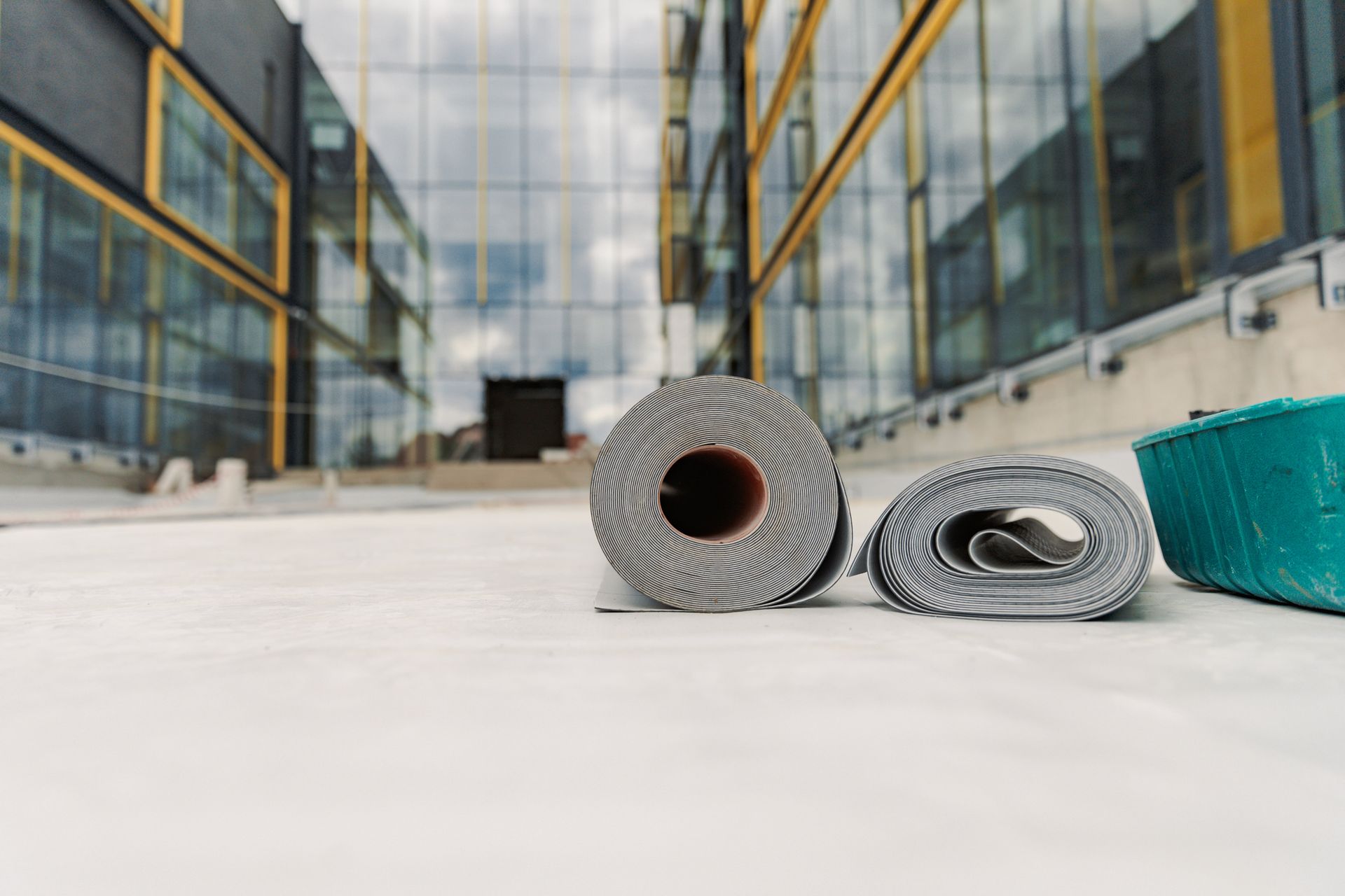 Two rolls of gray roofing material on a concrete surface, with a modern building in the background.