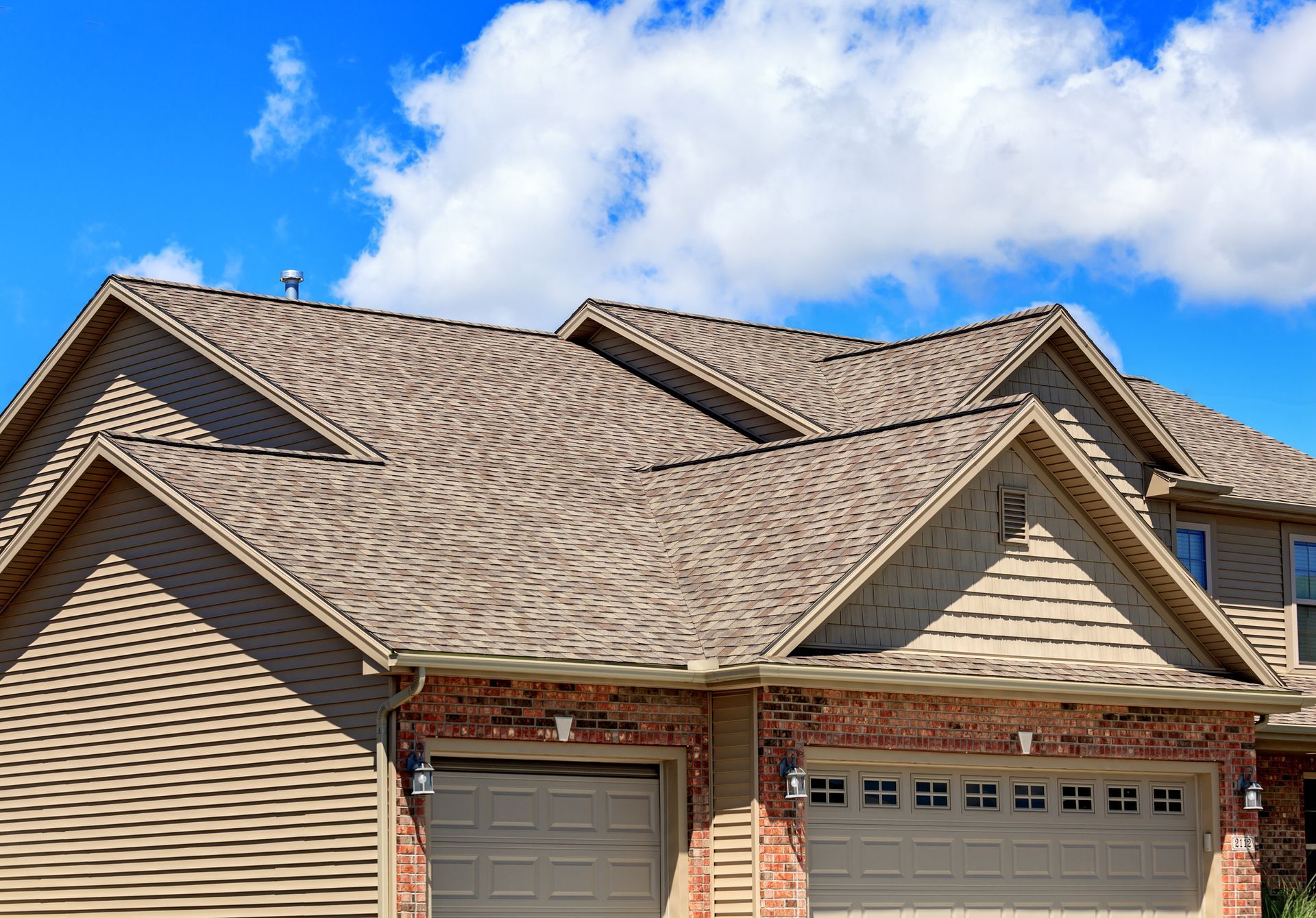 Tan shingled roof on a two-story house with a blue sky and clouds.
