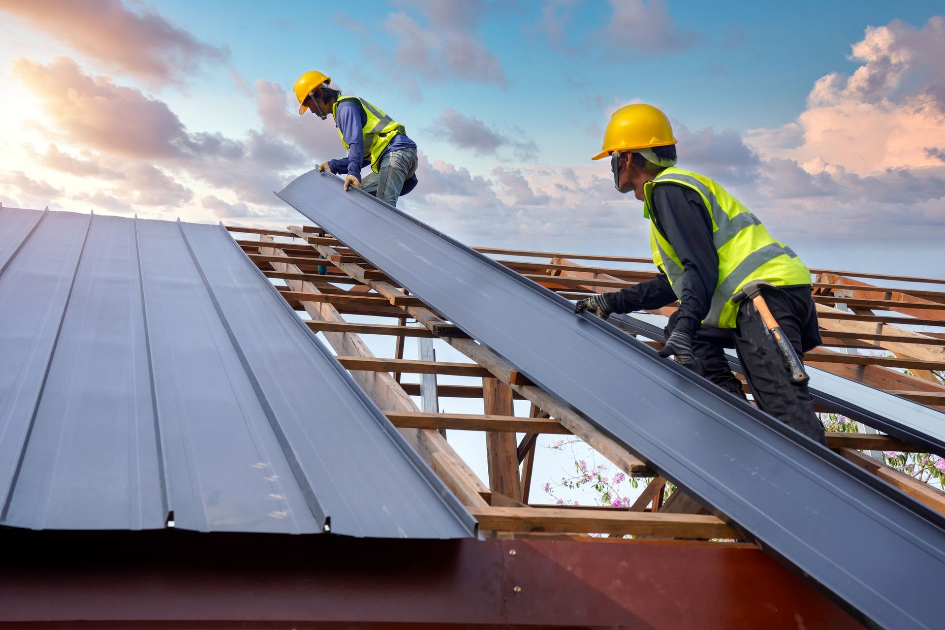 Two roofers in yellow helmets install metal roofing on a house frame against a cloudy sky.