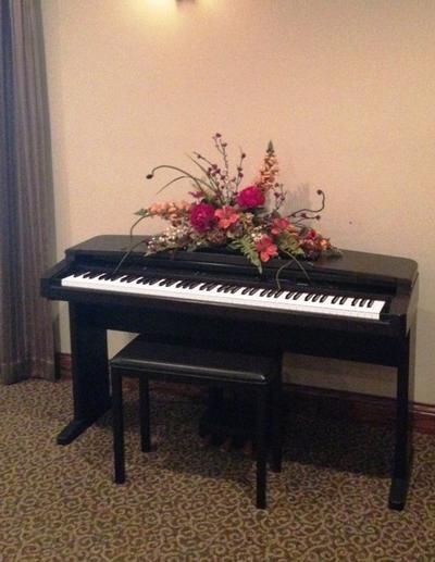 Black digital piano with a floral arrangement on top against a beige wall, with a small bench in front.