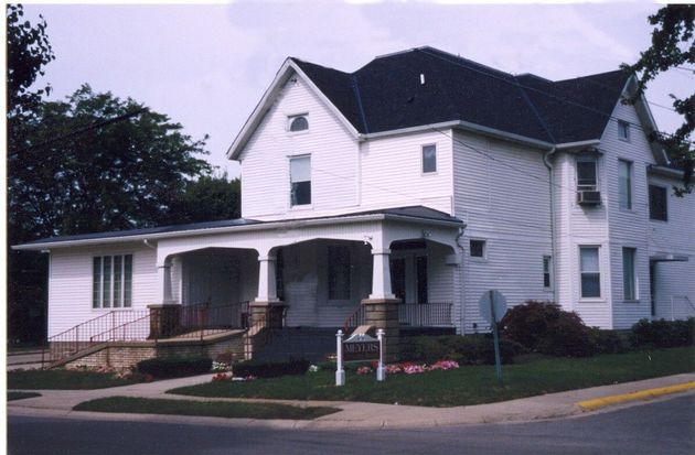 White two-story house with a covered porch and a small front yard on a street corner