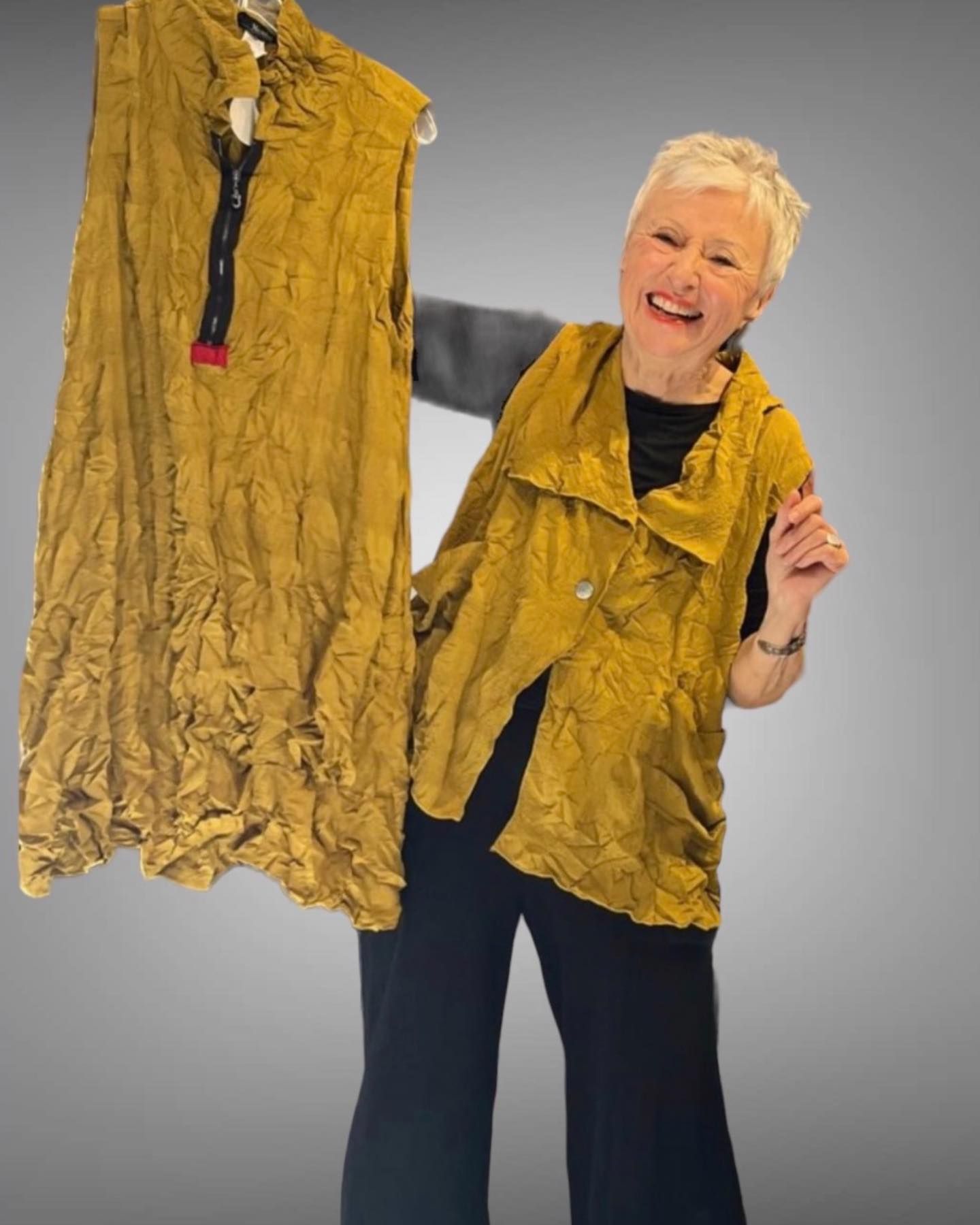 Woman holding up two mustard-yellow garments, smiling; standing in front of a gray backdrop.