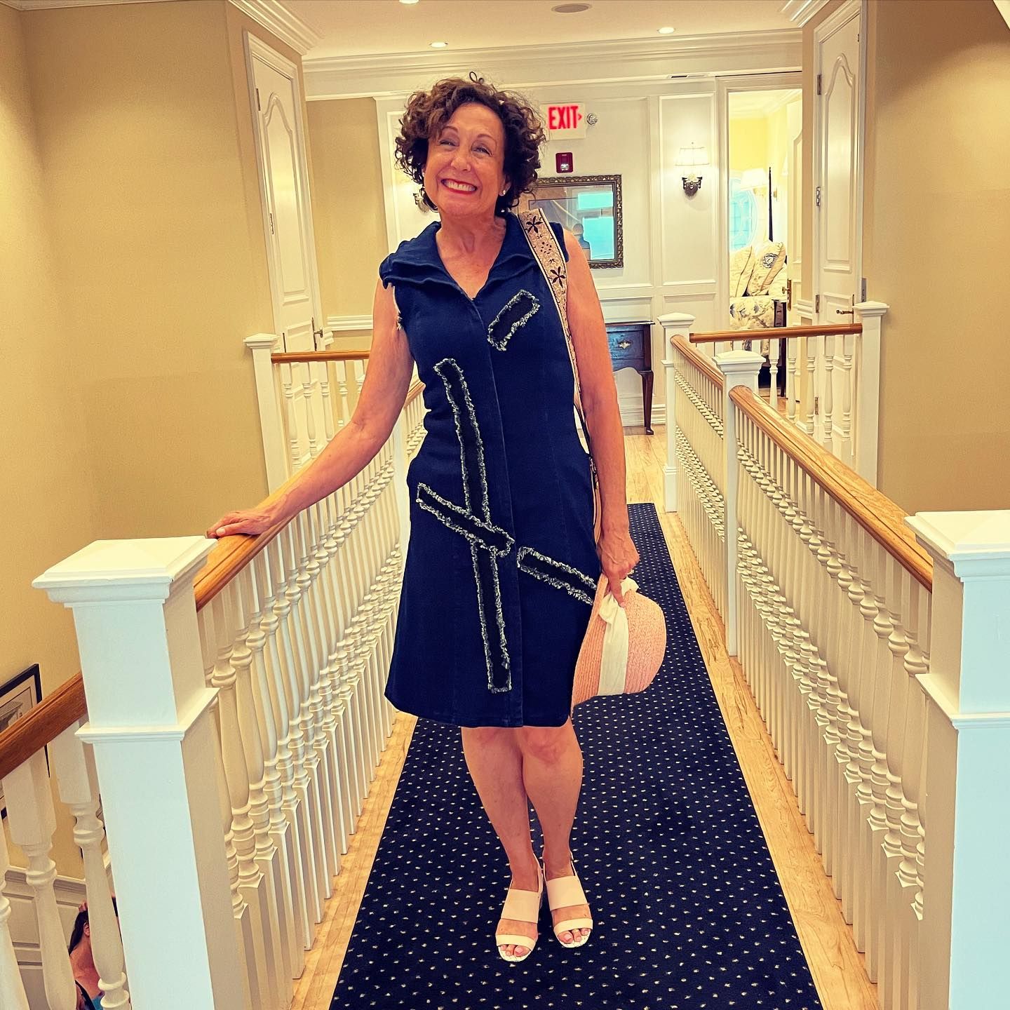 Woman in blue dress stands on a stairwell, holding a purse and hat.