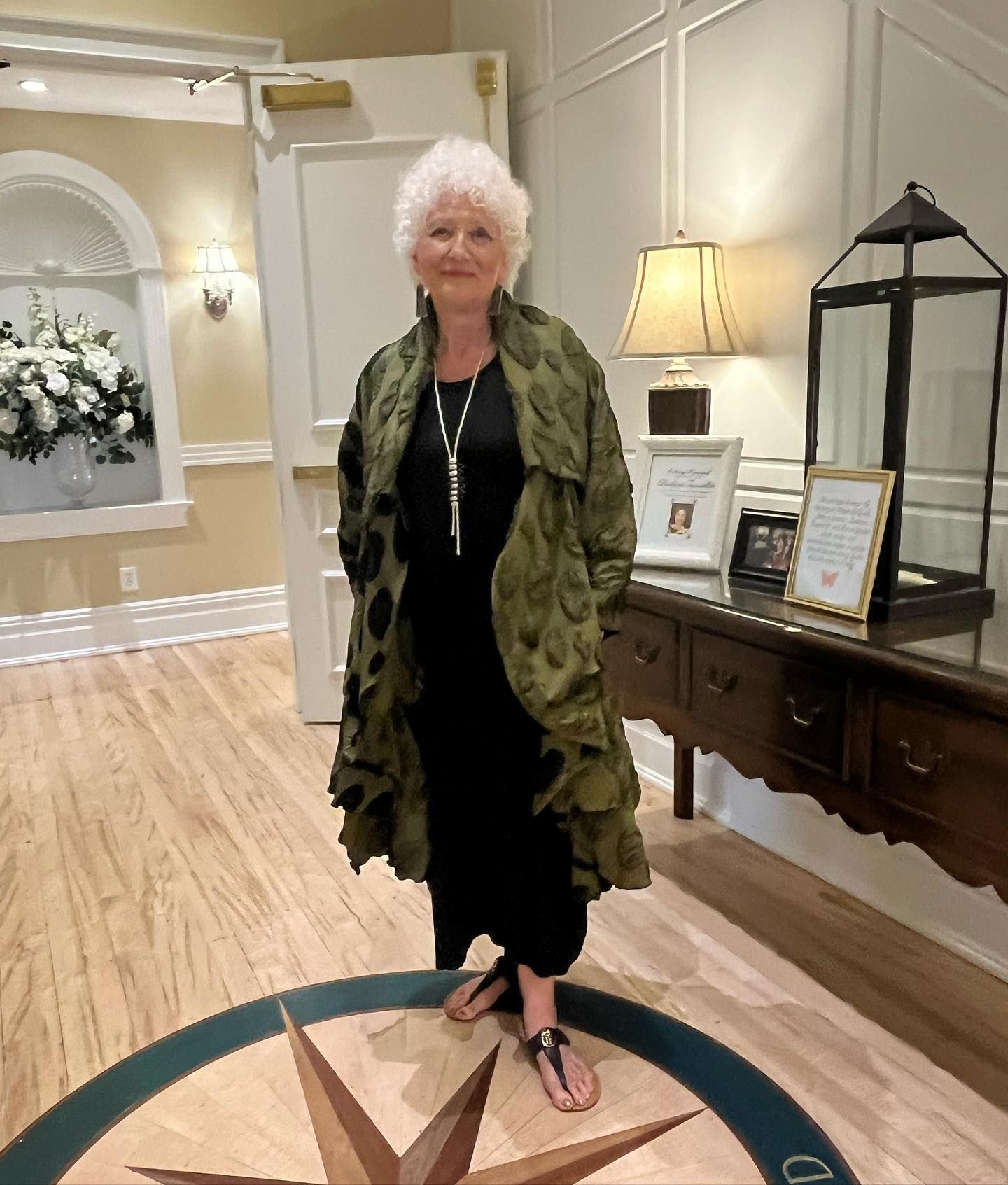 Woman in green patterned coat and black dress stands indoors near a decorative floor inlay, a table, and a door.