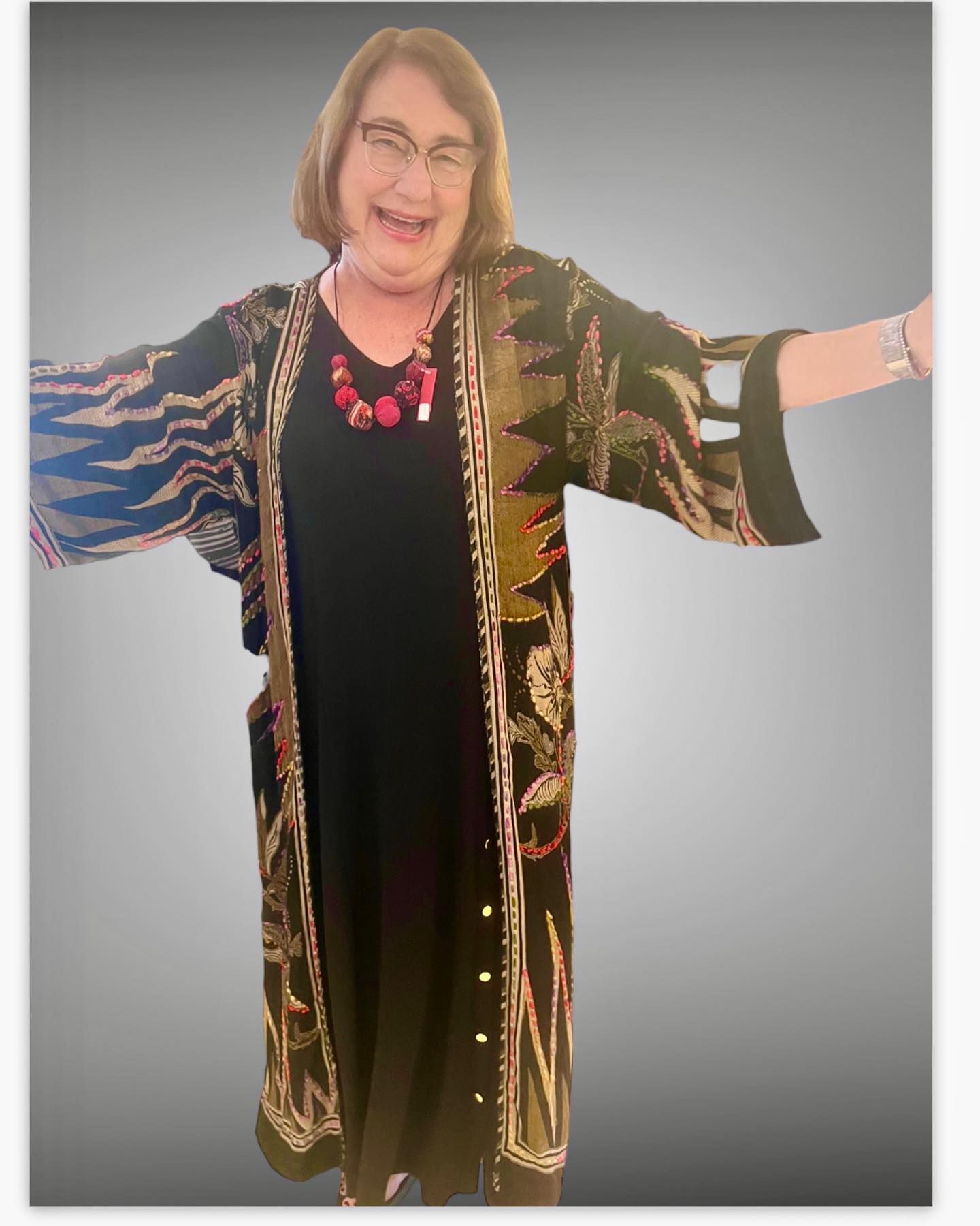 Woman with glasses smiles, arms outstretched, wearing a patterned kimono over a black dress.