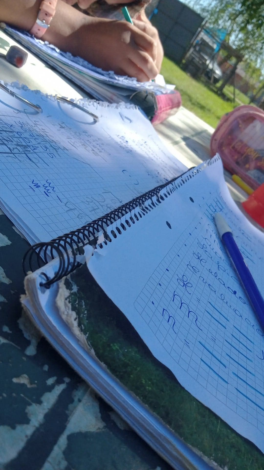 Cuadernos con notas escritas a mano y un lápiz azul sobre una mesa al aire libre; persona escribiendo en el fondo.