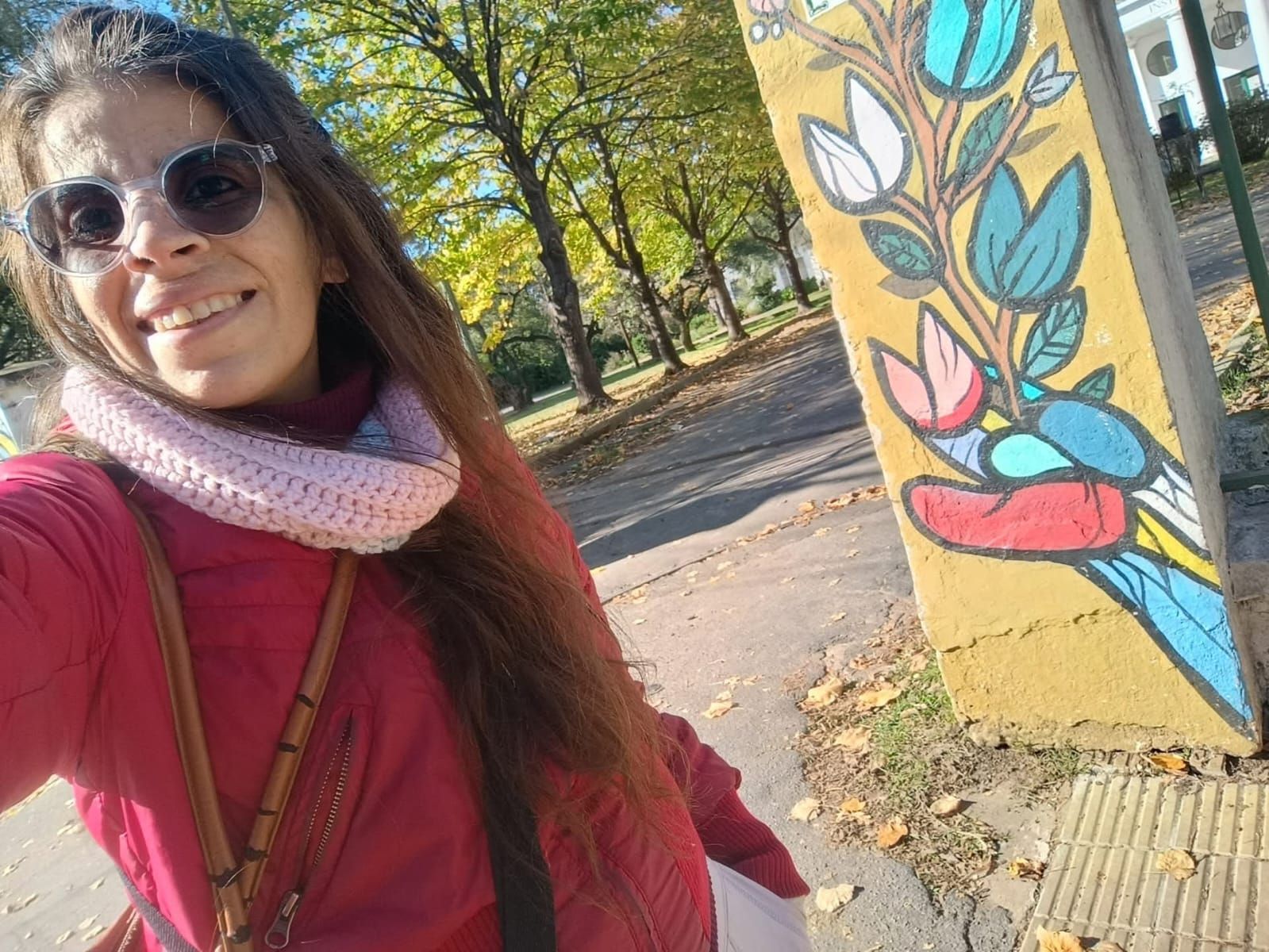 Una mujer con gafas de sol sonríe junto a un colorido mural al aire libre en un día soleado.