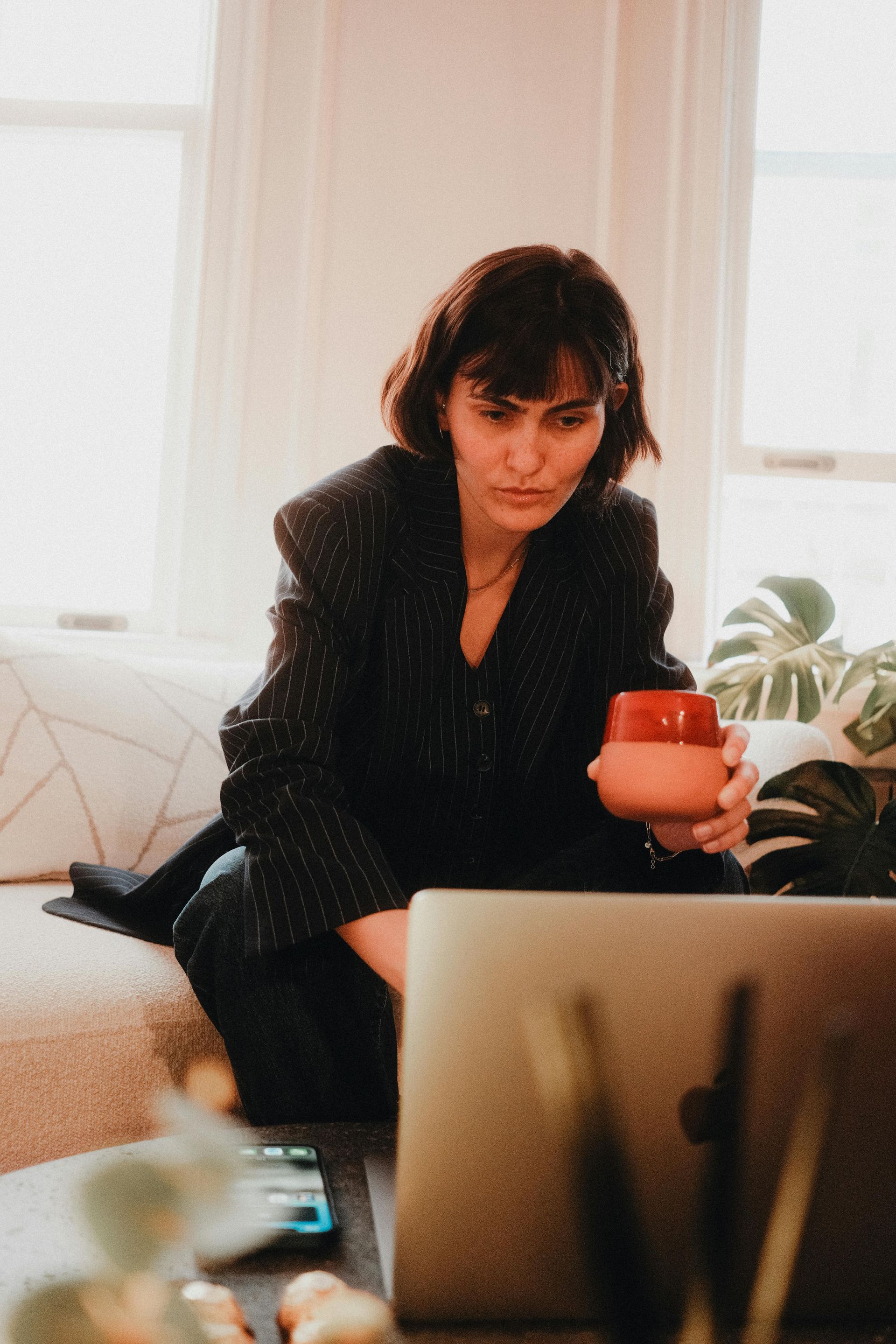 Mujer con blazer, sosteniendo una taza, mirando fijamente la pantalla de un portátil.