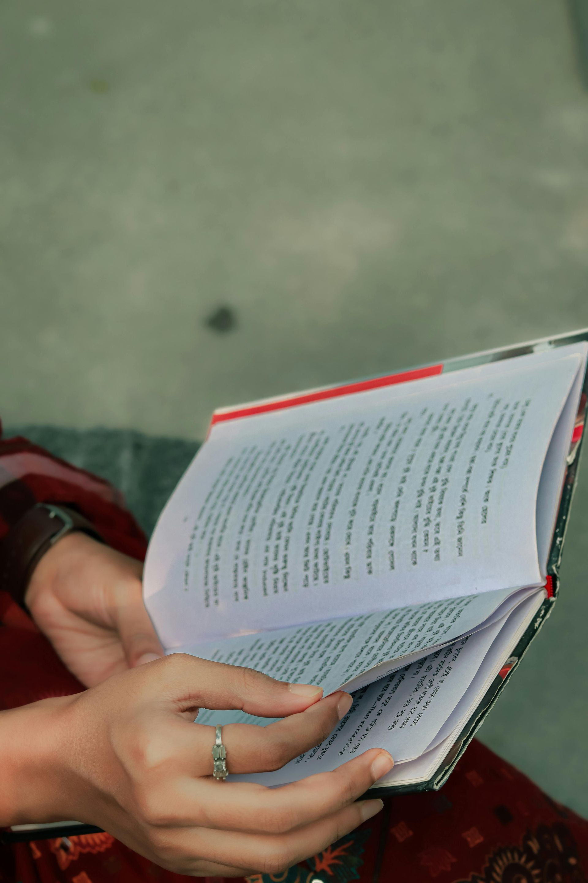 Manos pasando las páginas de un libro; tapa roja, anillo en el dedo, sentado al aire libre.