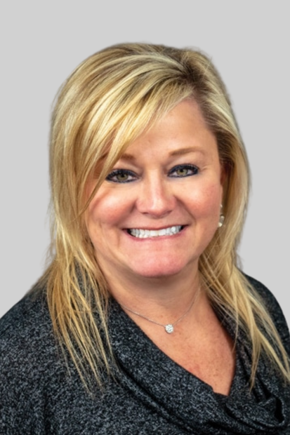 Smiling man in a light blue shirt standing in front of a wall with a partial logo.