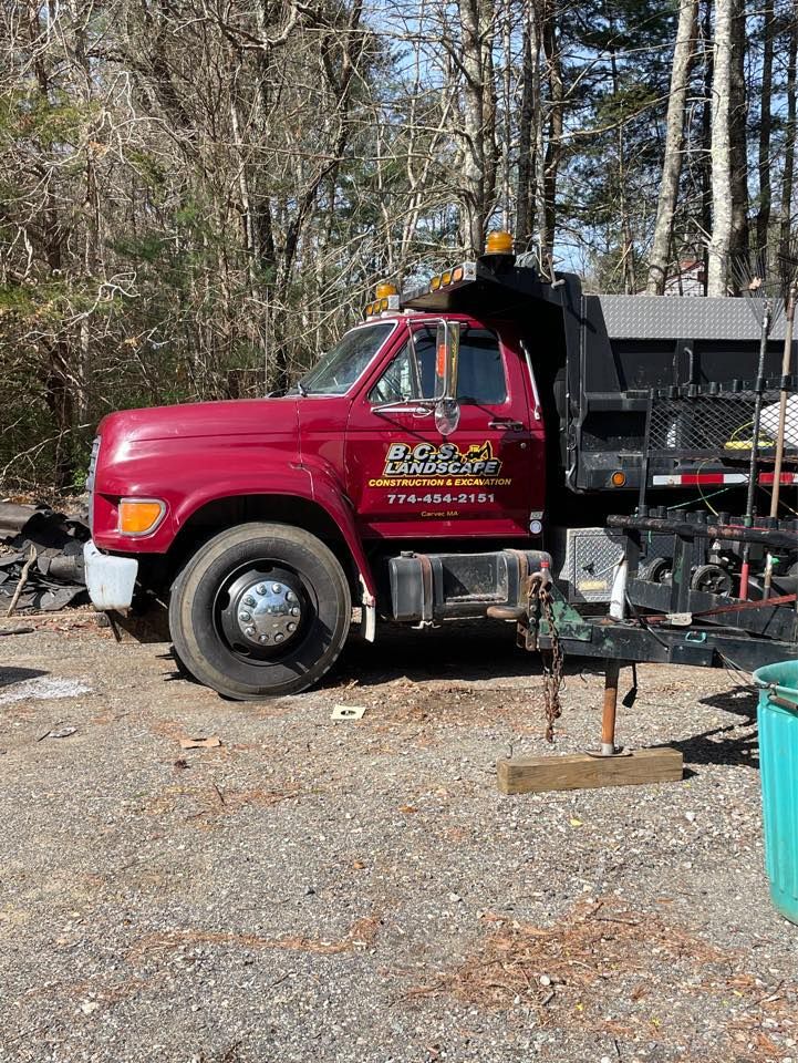 Red dump truck parked on gravel, with company name on the door and trees in the background.