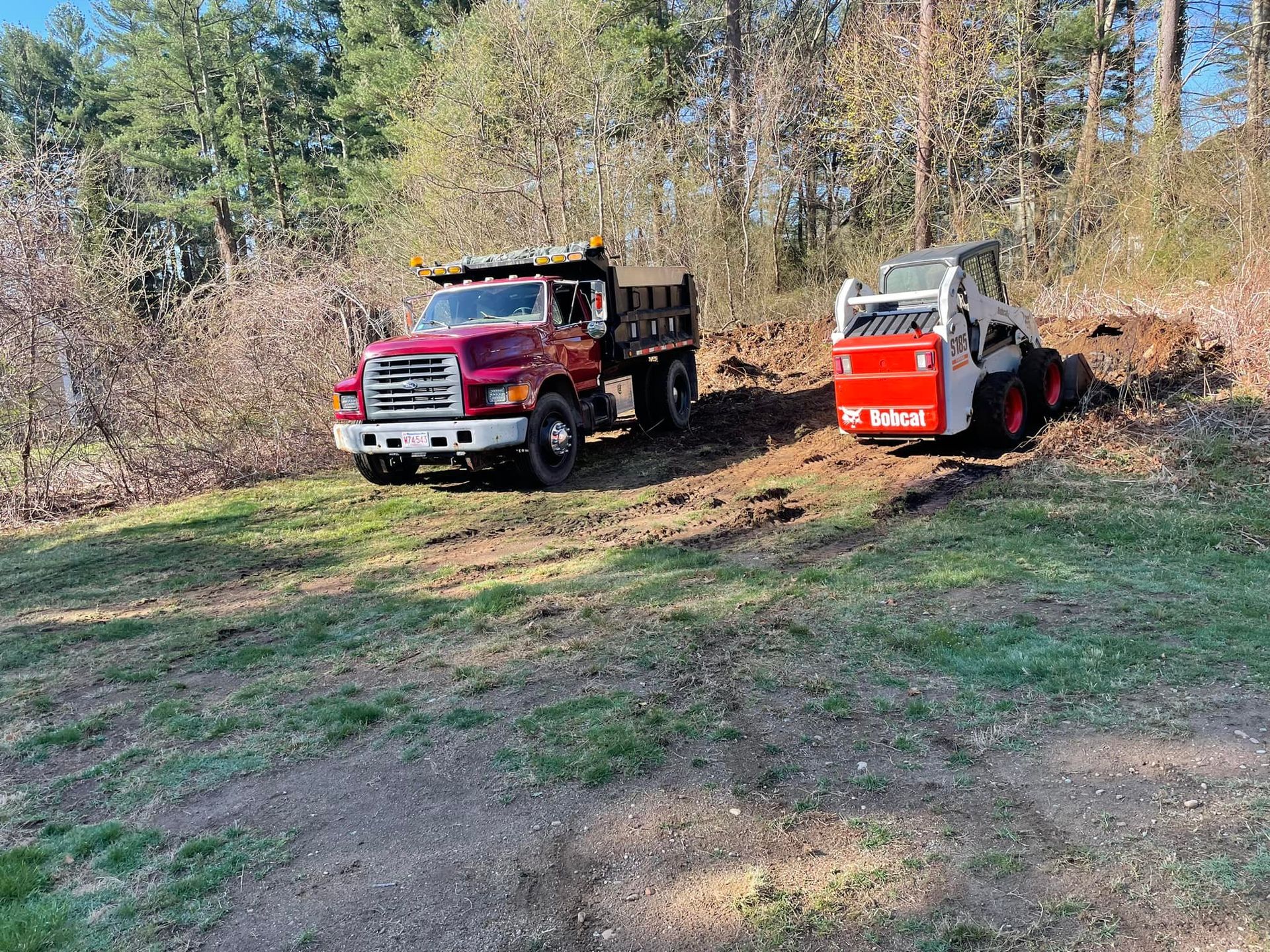 A red dump truck and a white Bobcat on a grassy hill, trees in the background.