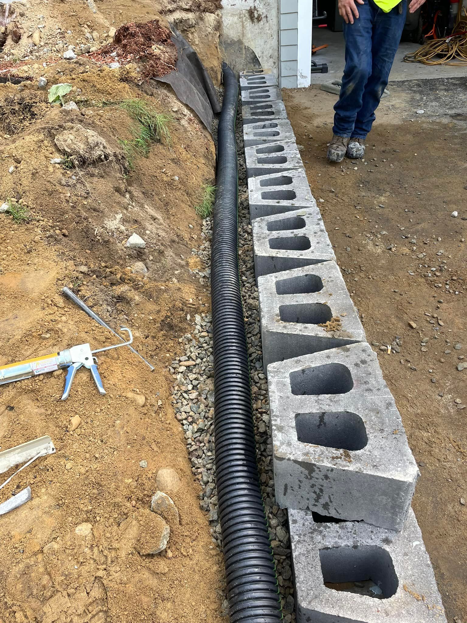 Black corrugated pipe beside a cinder block wall, set in gravel. Person in jeans standing nearby.
