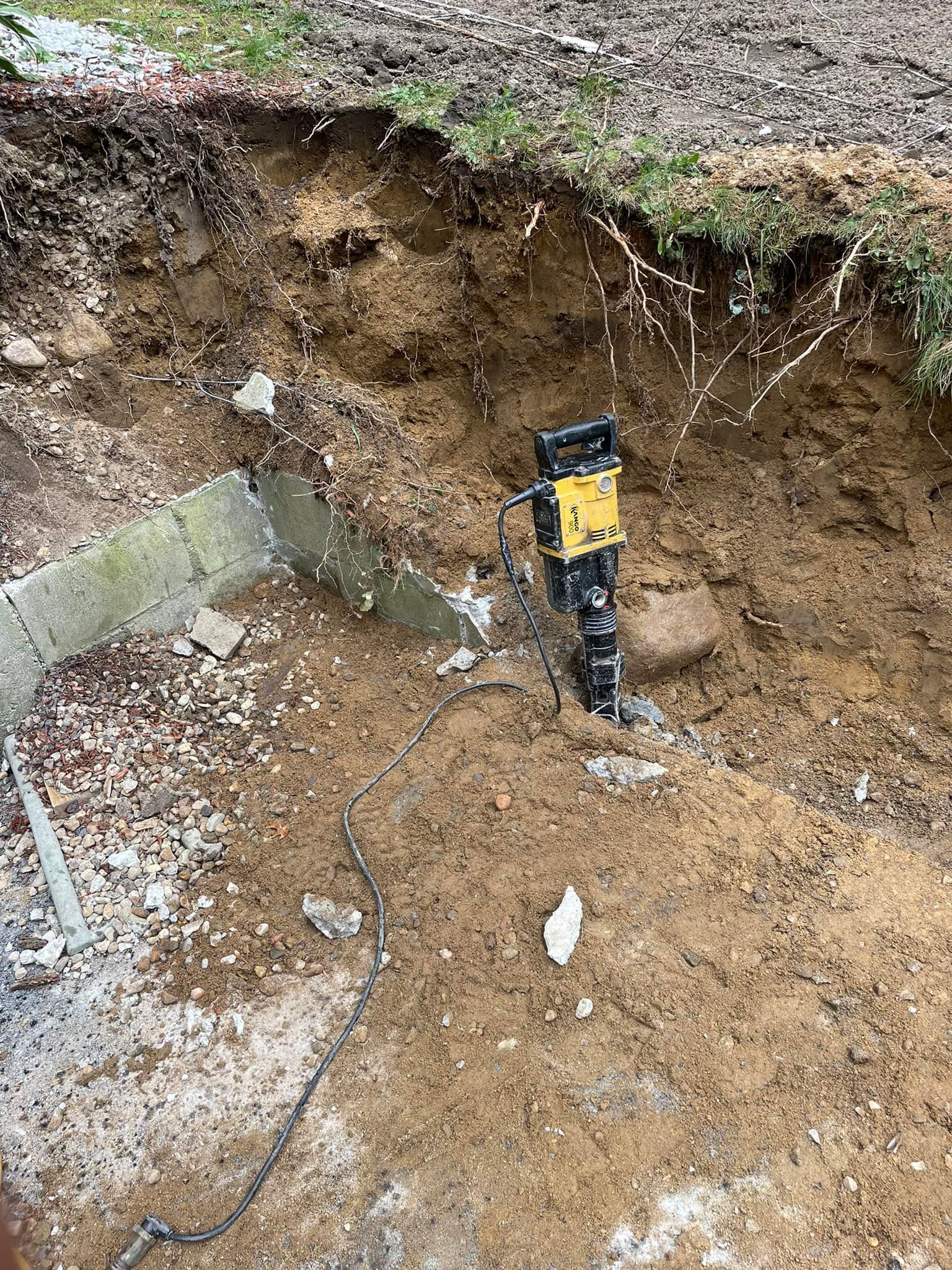A jackhammer in a dug-out area, with a metal structure, soil, and grass visible.