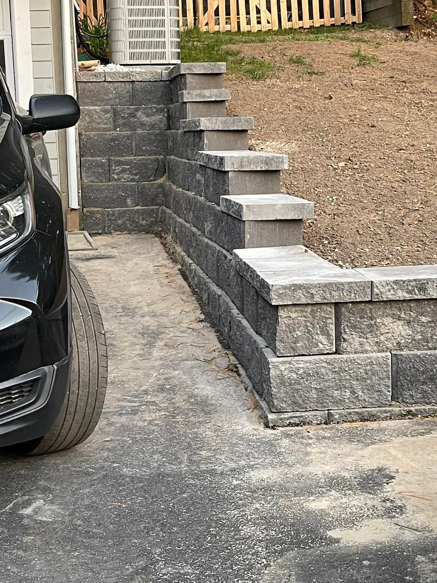 Black car next to concrete steps and retaining wall leading to a grassy hill.