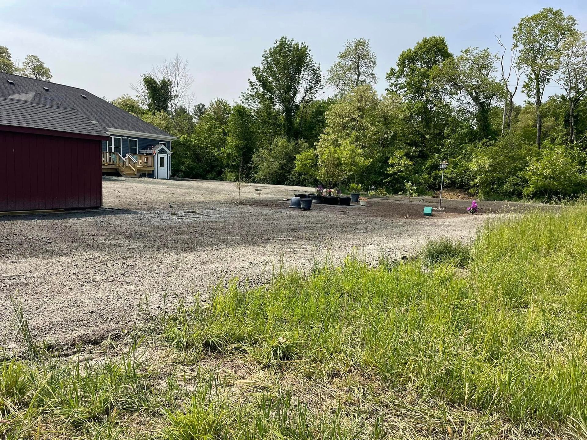 Gravel lot with house and trees in the background. Green grass in the foreground under a cloudy sky.