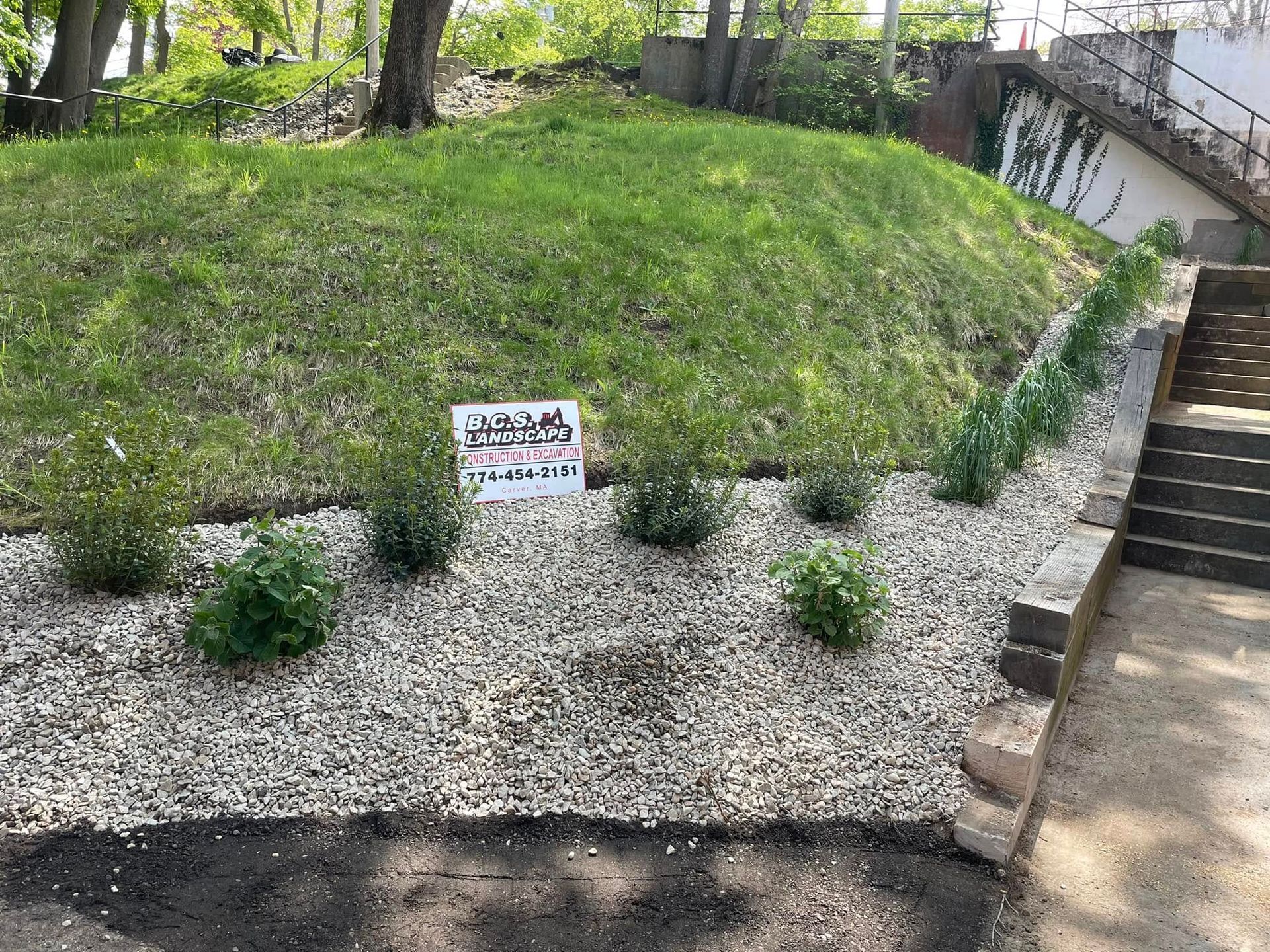 Landscaped yard with small green bushes, gravel ground cover, and a grassy hill in the background.