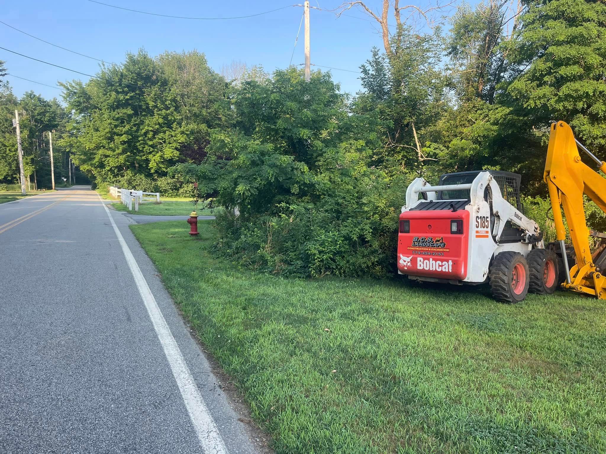 A red and white Bobcat skid steer next to a roadside clearing overgrown with bushes.